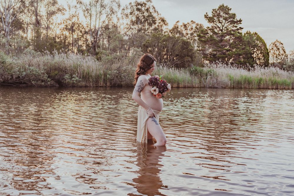 Expecting mother in white dress holding flowers while standing in serene water.