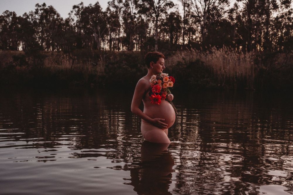 Expecting mother stands in still water at dusk holding red flowers.