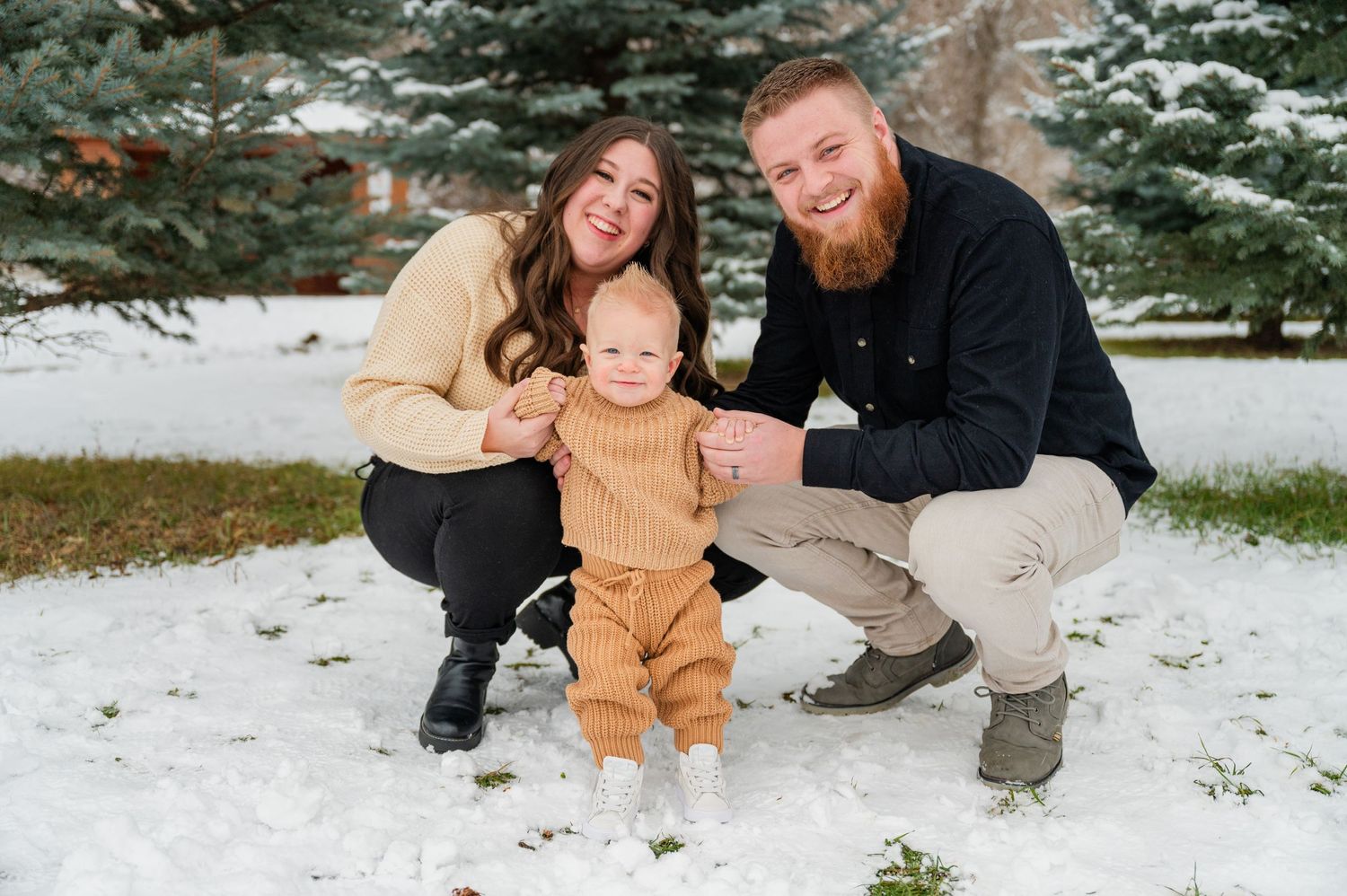 A family of three poses together in a snowy setting surrounded by evergreen trees.
