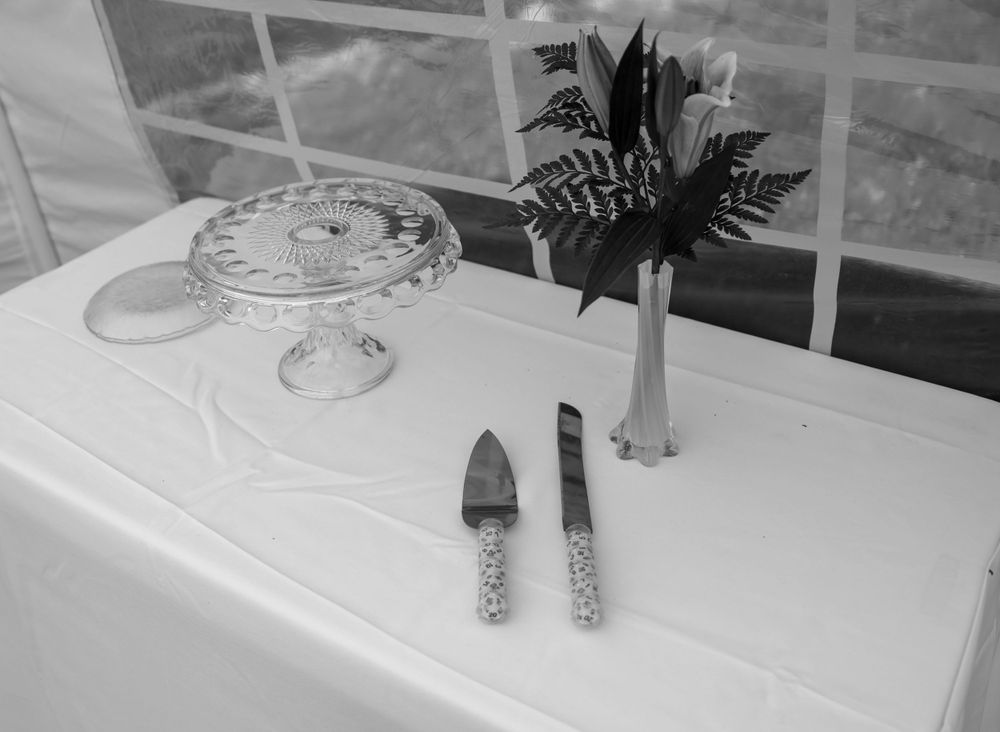 Black and white photo of cake serving set beside small flower arrangement on white tablecloth.