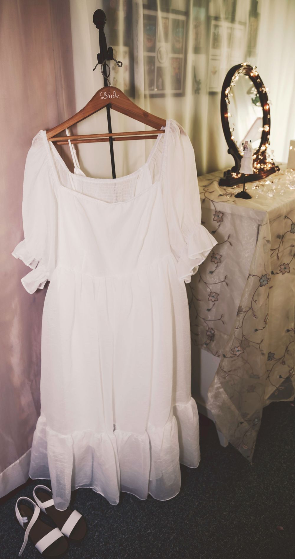 A white vintage dress with puff sleeves hangs on a wooden hanger beside an ornate mirror and white curtains.