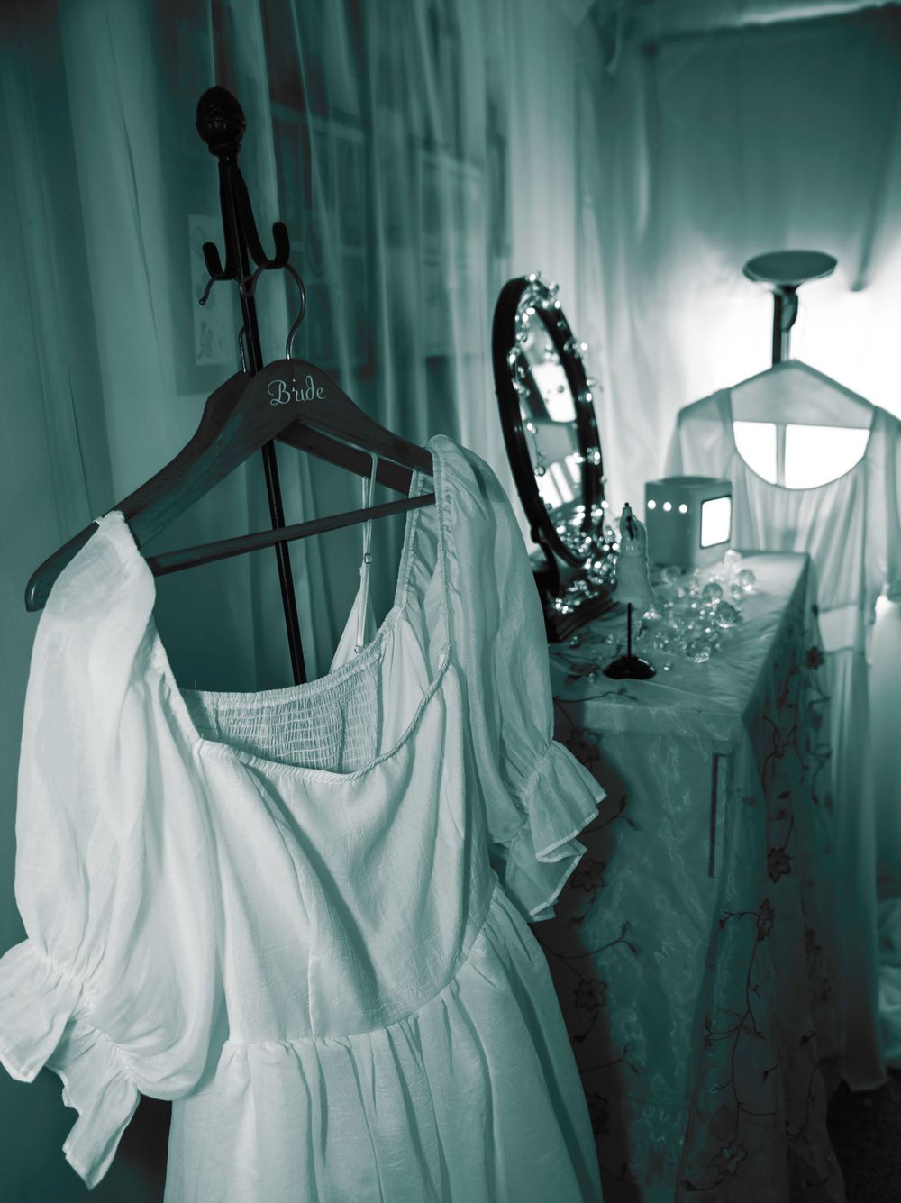Ethereal white dress displayed on hanger with antique mirror in a moody teal-tinted setting.