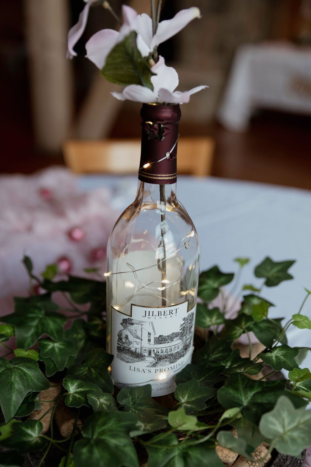 White flowers arranged in a vintage wine bottle centerpiece with green ivy leaves on a table.