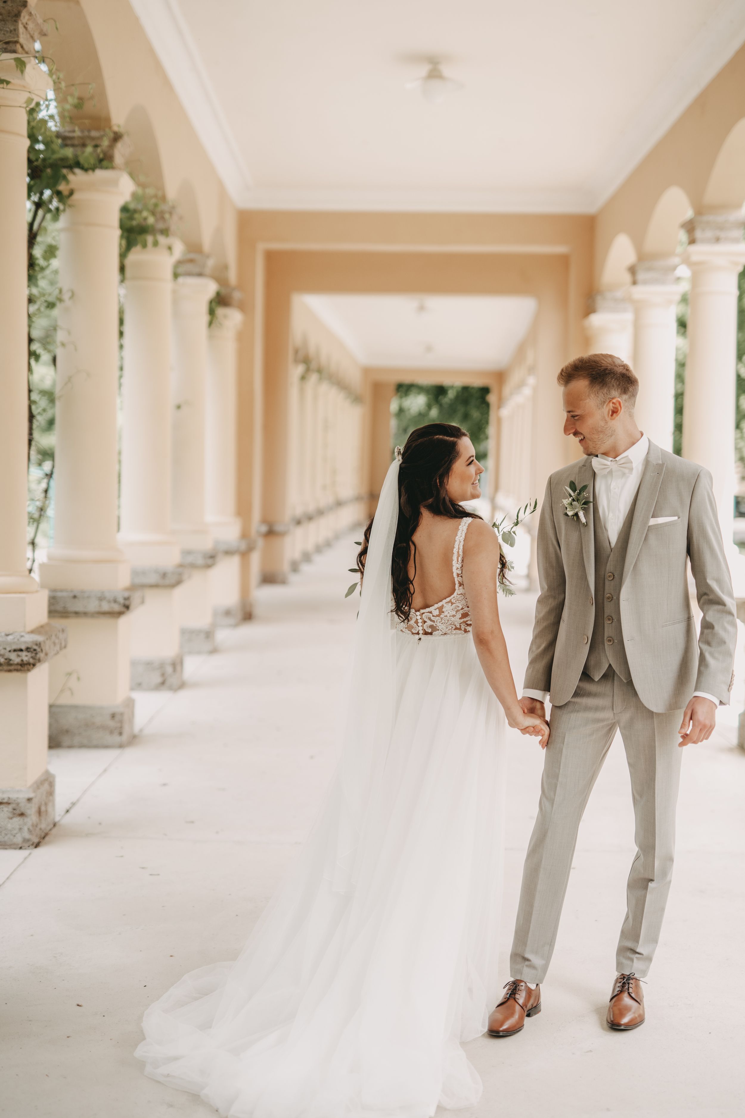 Elegant couple in wedding attire standing in classical colonnade with decorative greenery.