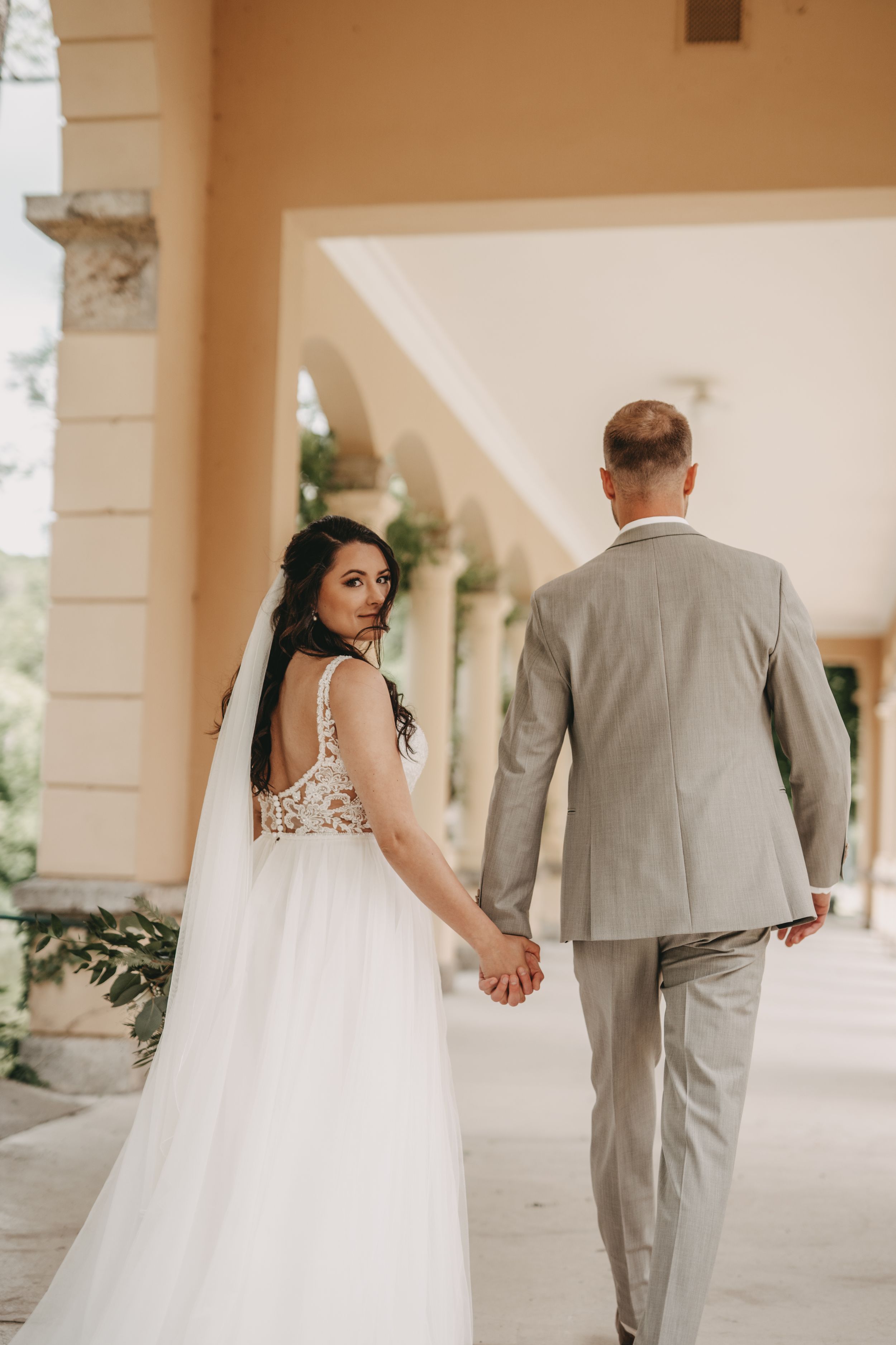 Bride in lace-back dress holds hands with groom while walking through columned hallway.
