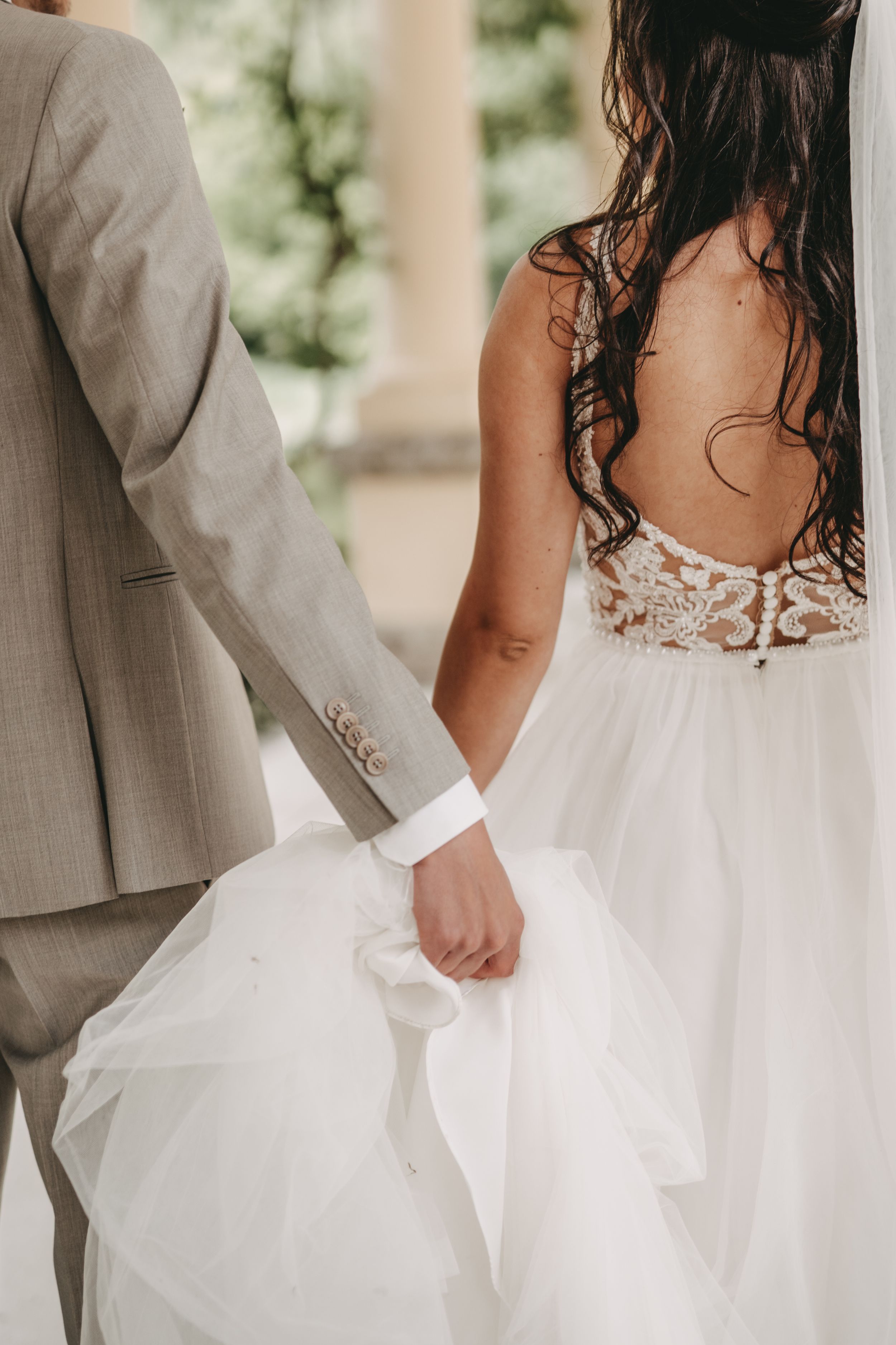 Close-up detail of a bride's lace back wedding dress and groom's gray suit as they stand together.