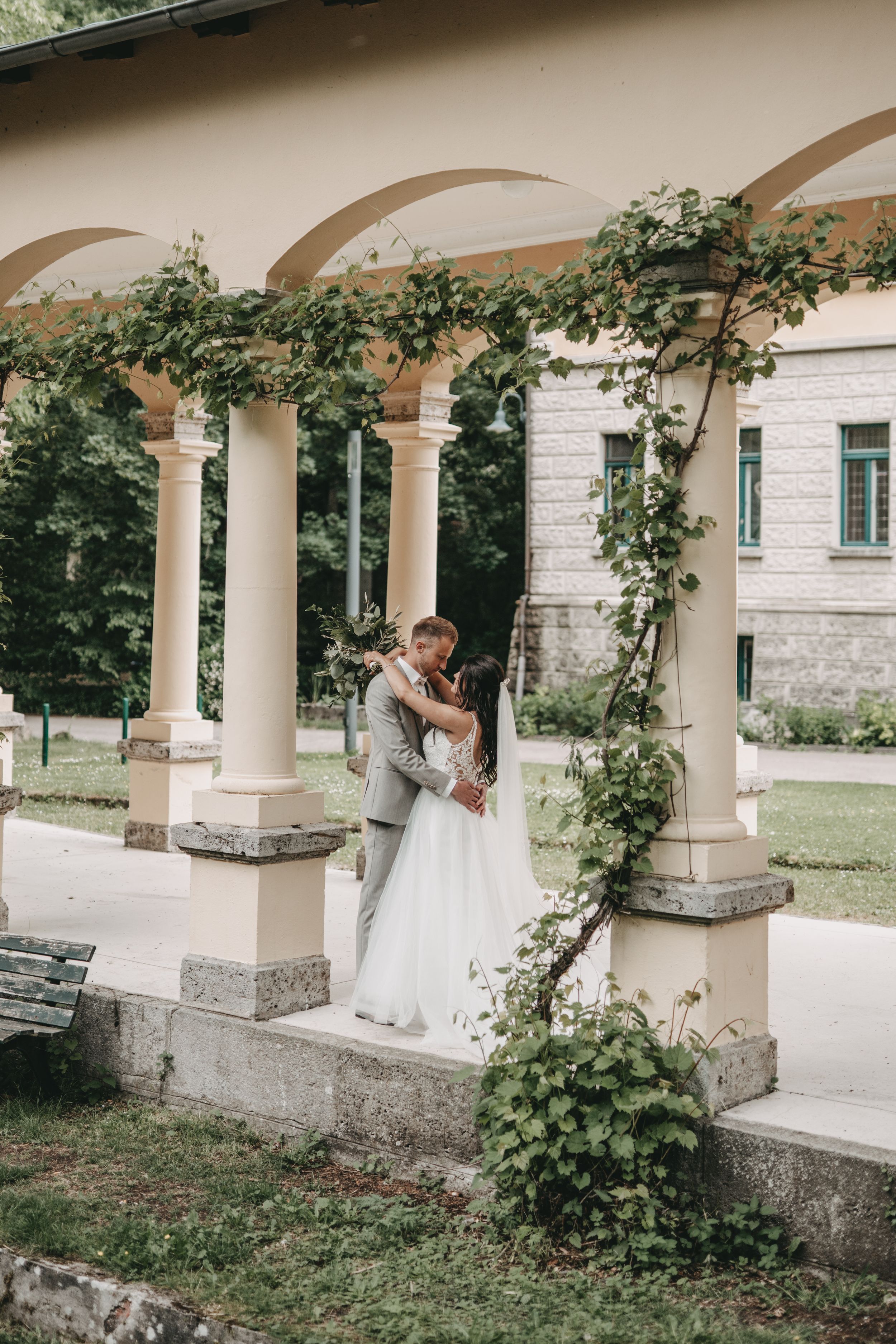 Wedding couple embracing under an elegant colonnade with climbing vines at a historic venue.