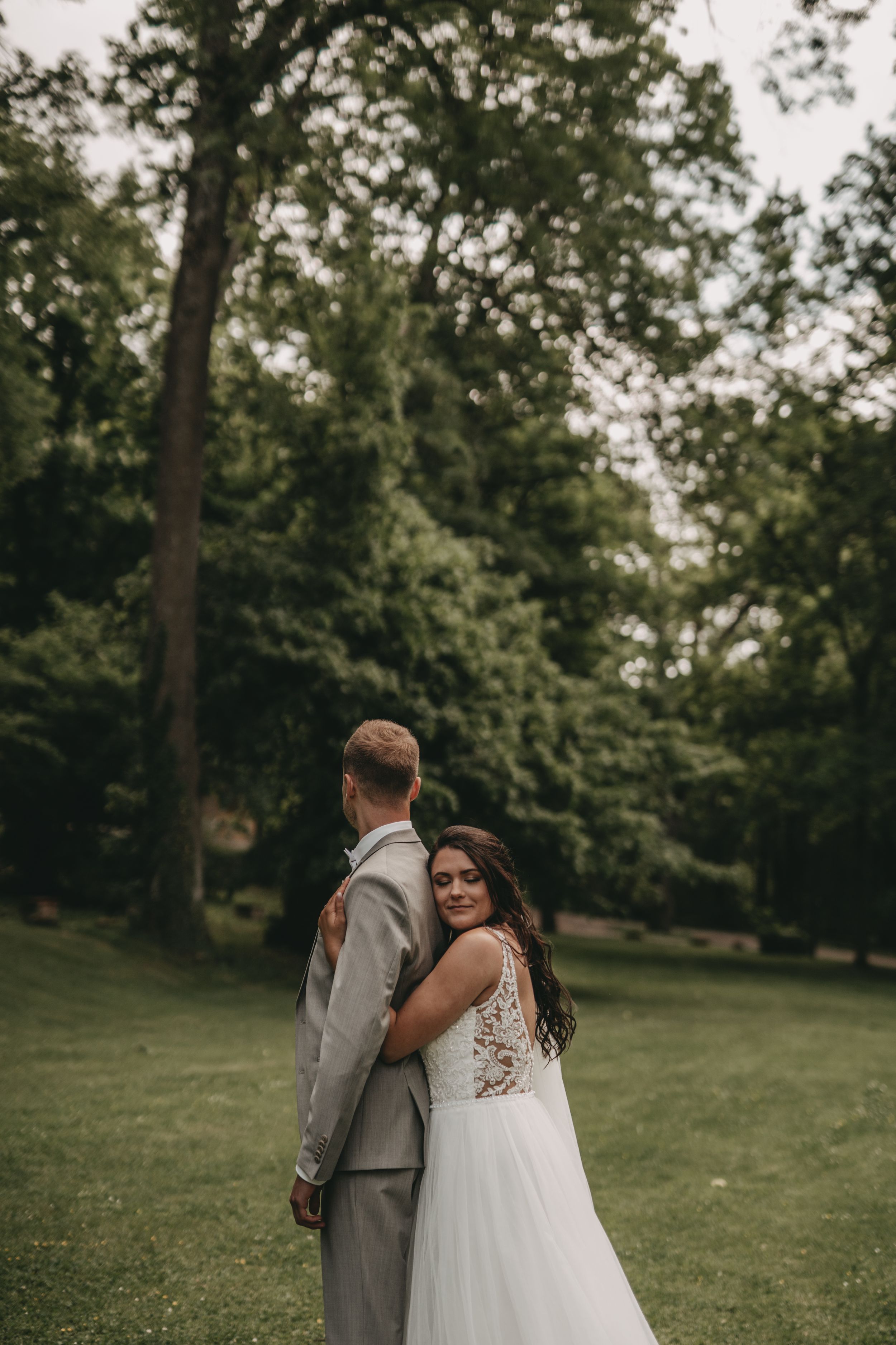 Newlyweds pose back-to-back on grassy lawn with trees in background.