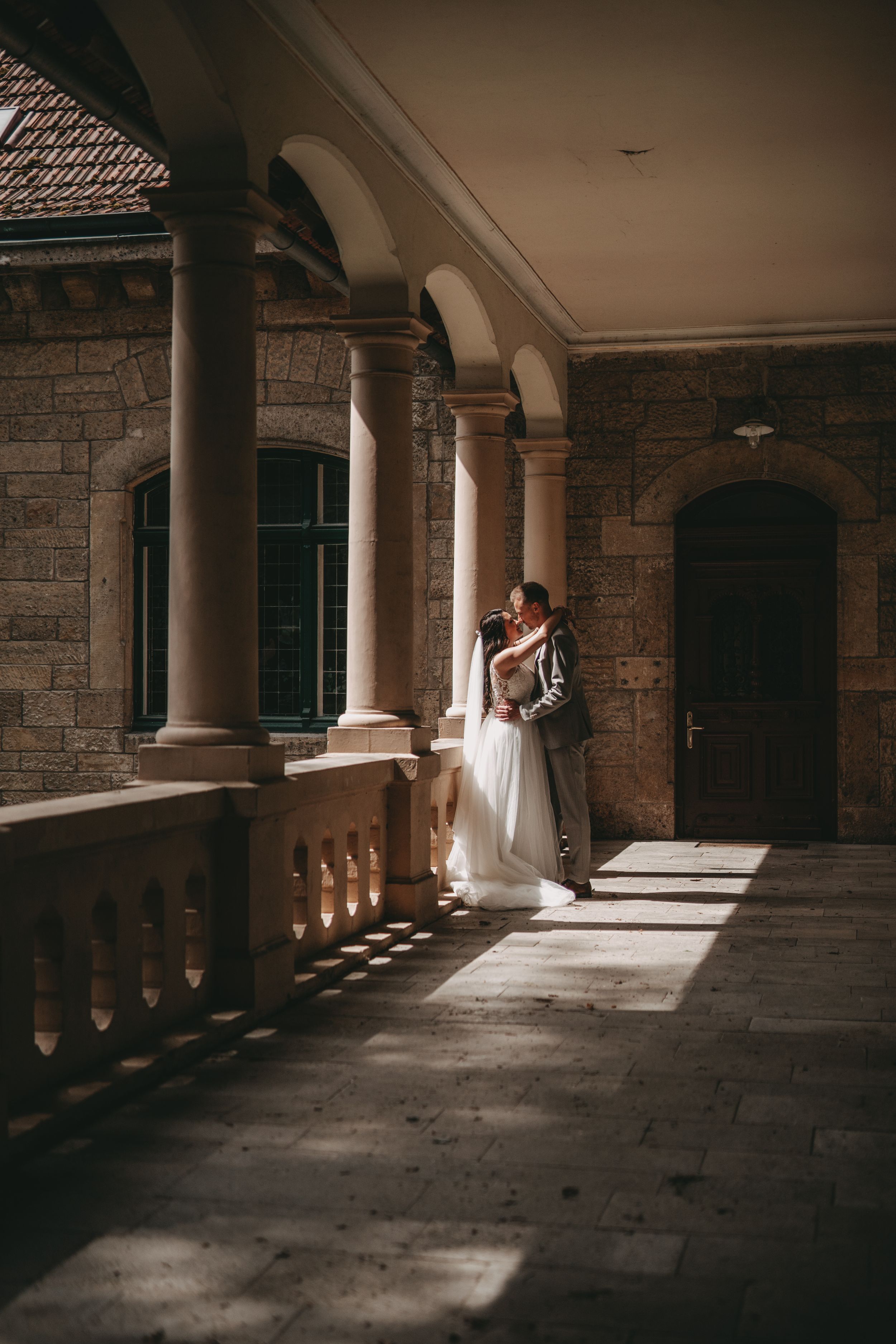 Wedding couple embracing in dramatic lighting under stone archway of historic building.
