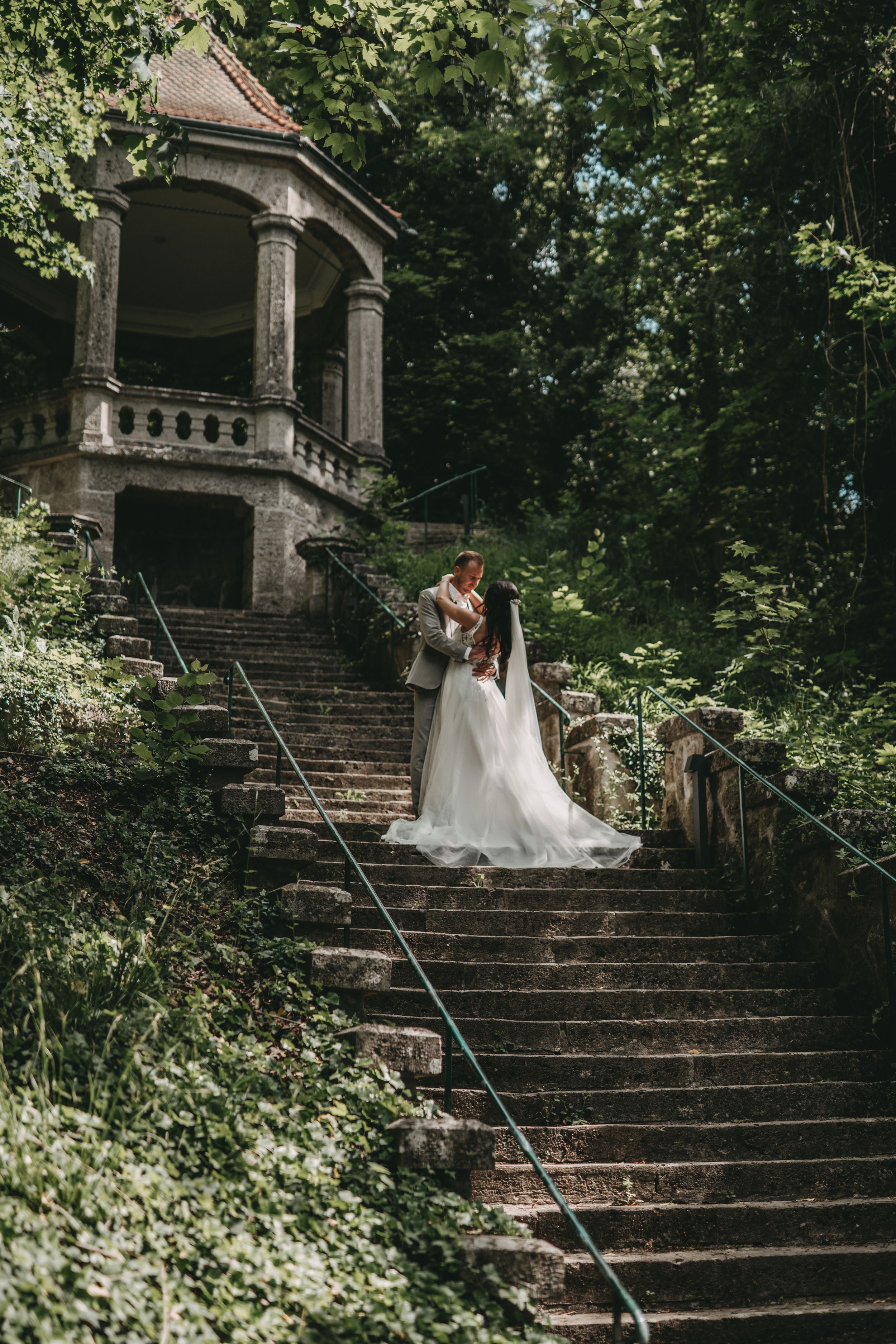 Wedding couple standing on stone steps leading to gazebo surrounded by lush greenery.