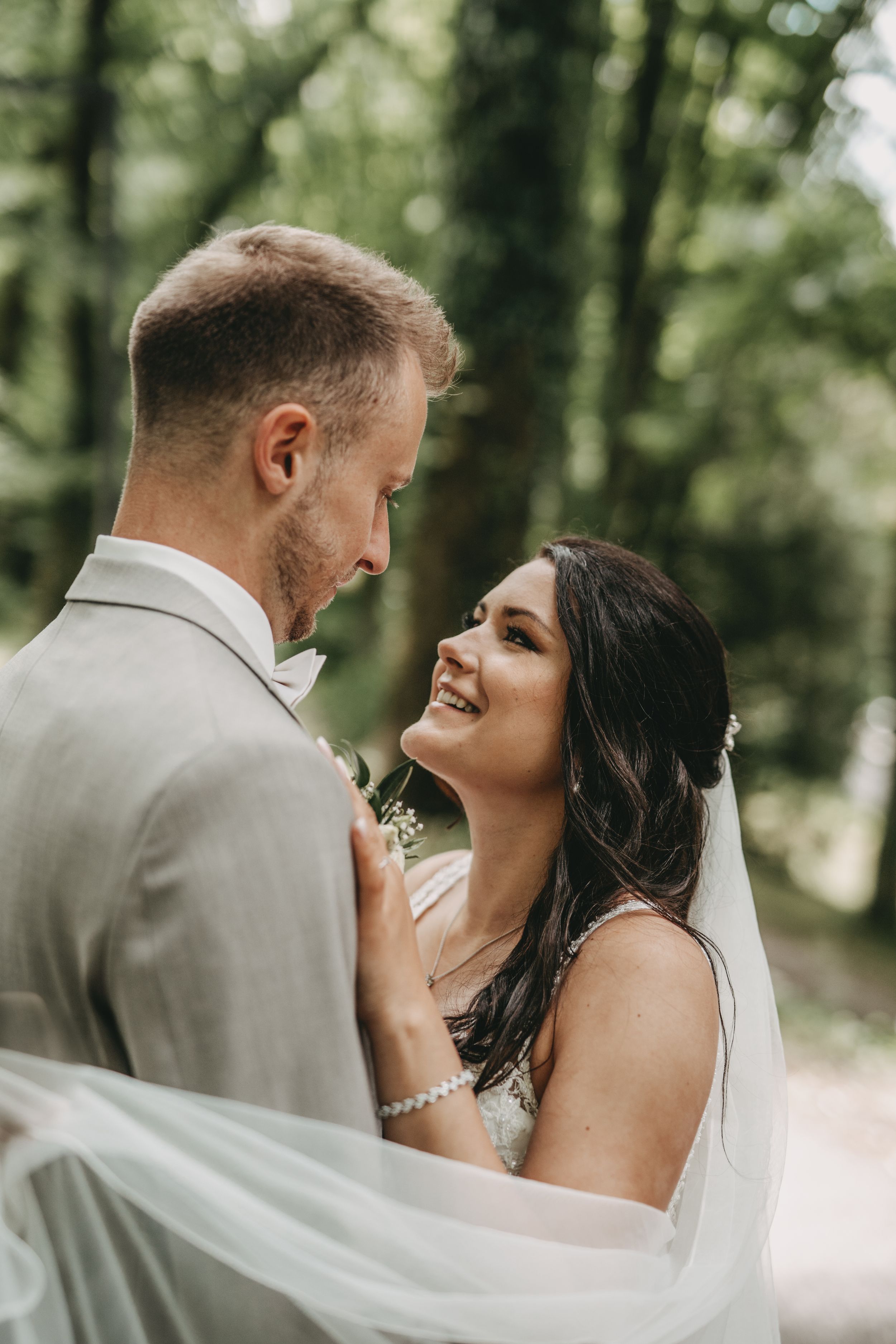 Romantic candid moment between newlyweds with flowing veil in forest setting.