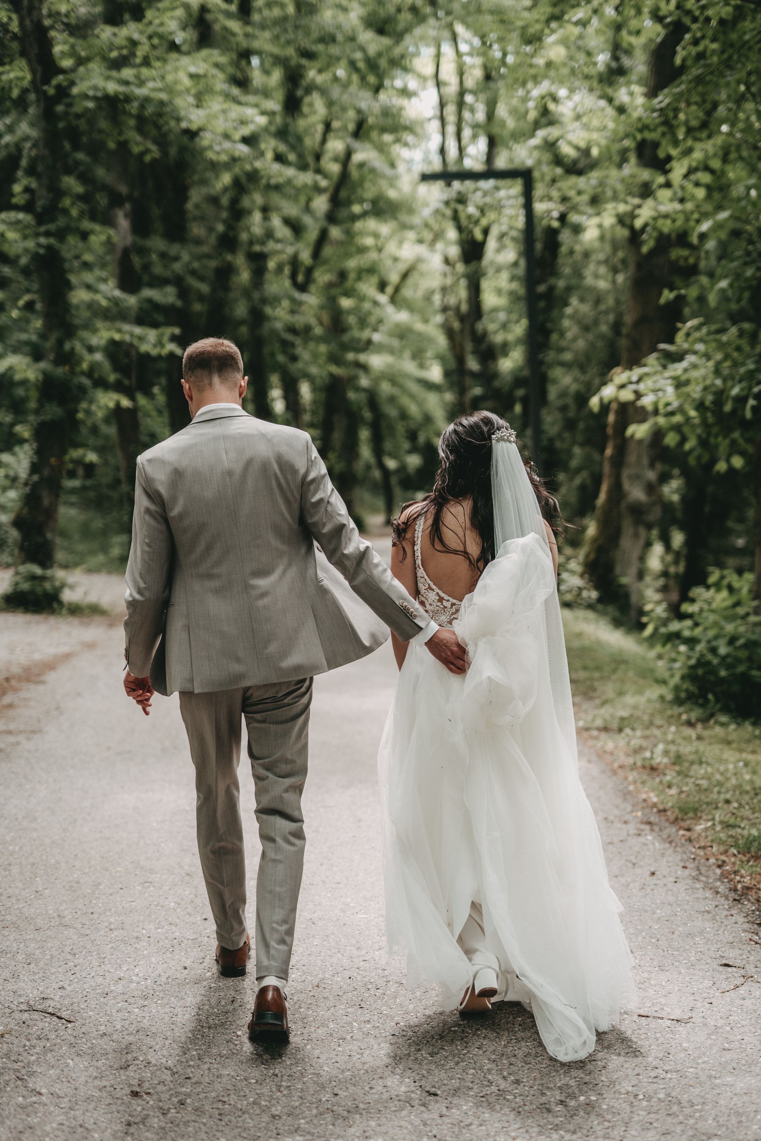 Newlyweds walking hand in hand down forest path in elegant wedding attire.
