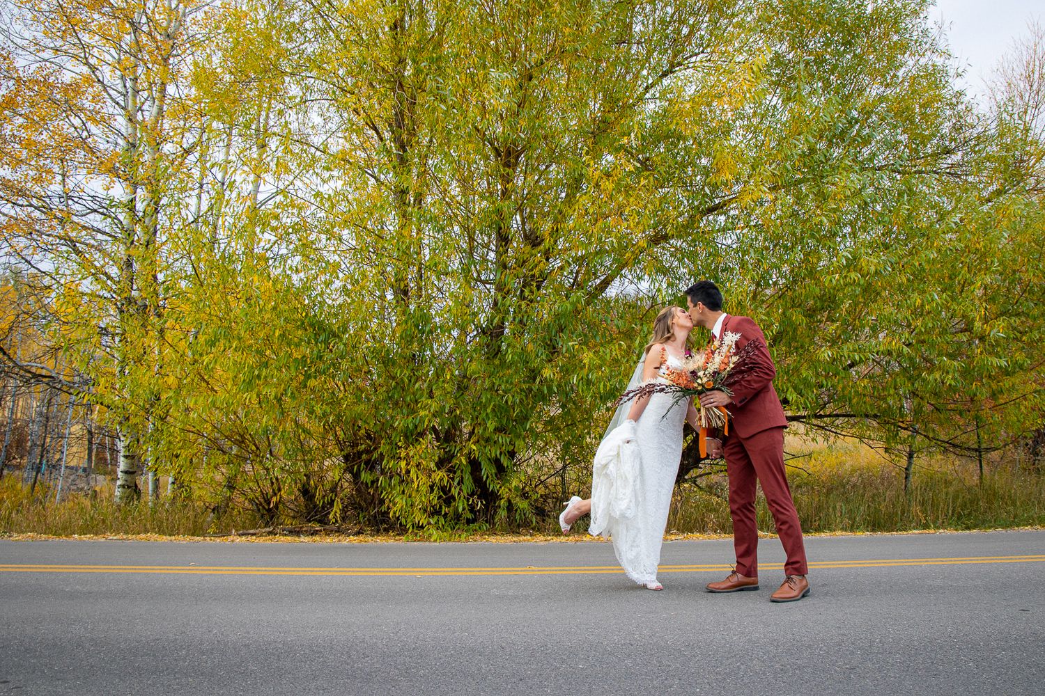 Romantic outdoor wedding couple embracing on autumn road with golden trees in background.