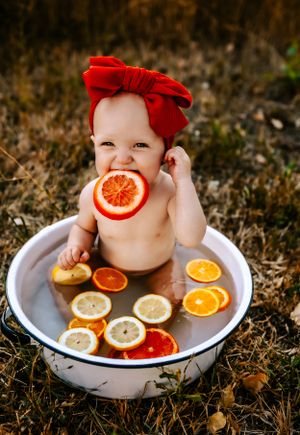 A baby wearing a red bow headband sits in a vintage tub with citrus slices for a creative photoshoot.