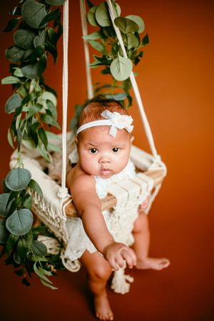 Baby sitting in macrame swing with eucalyptus garland against orange background.