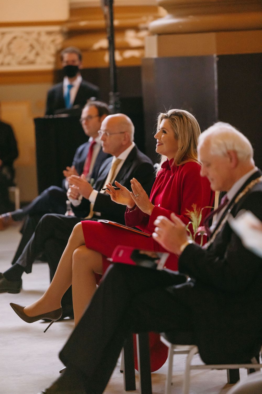 People seated in formal attire at an event with someone in a red dress gesturing while speaking.