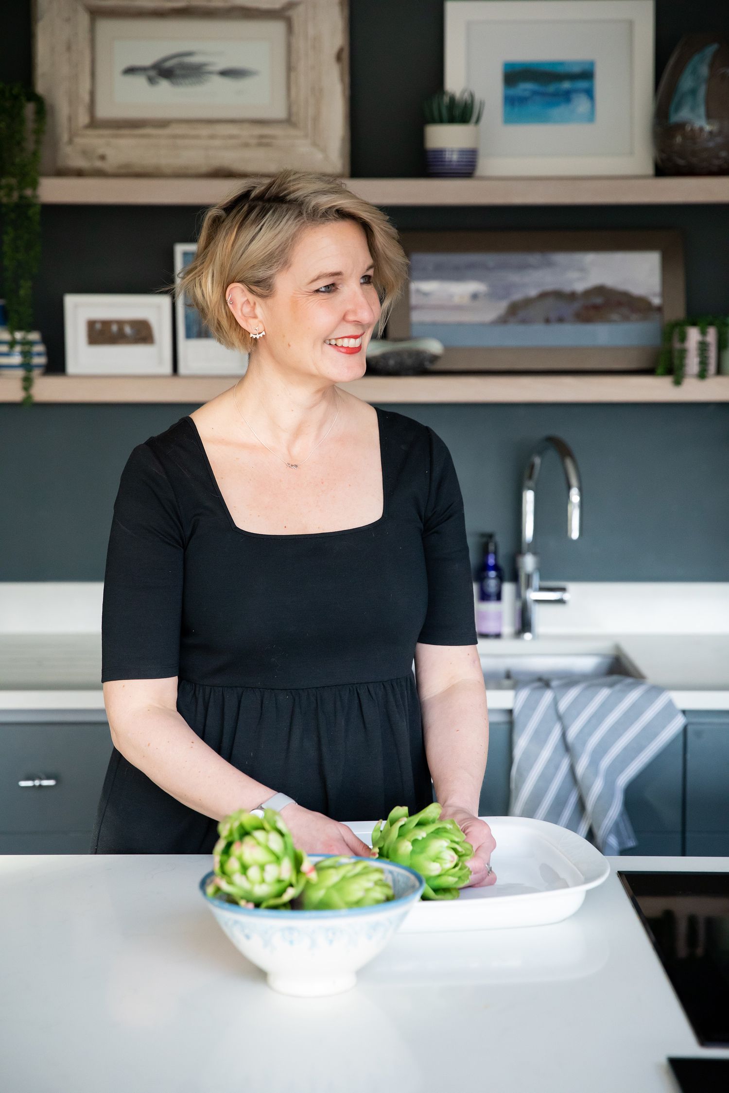 Person in black dress preparing artichokes in modern kitchen with framed artwork on wall during business branding photoshoot.