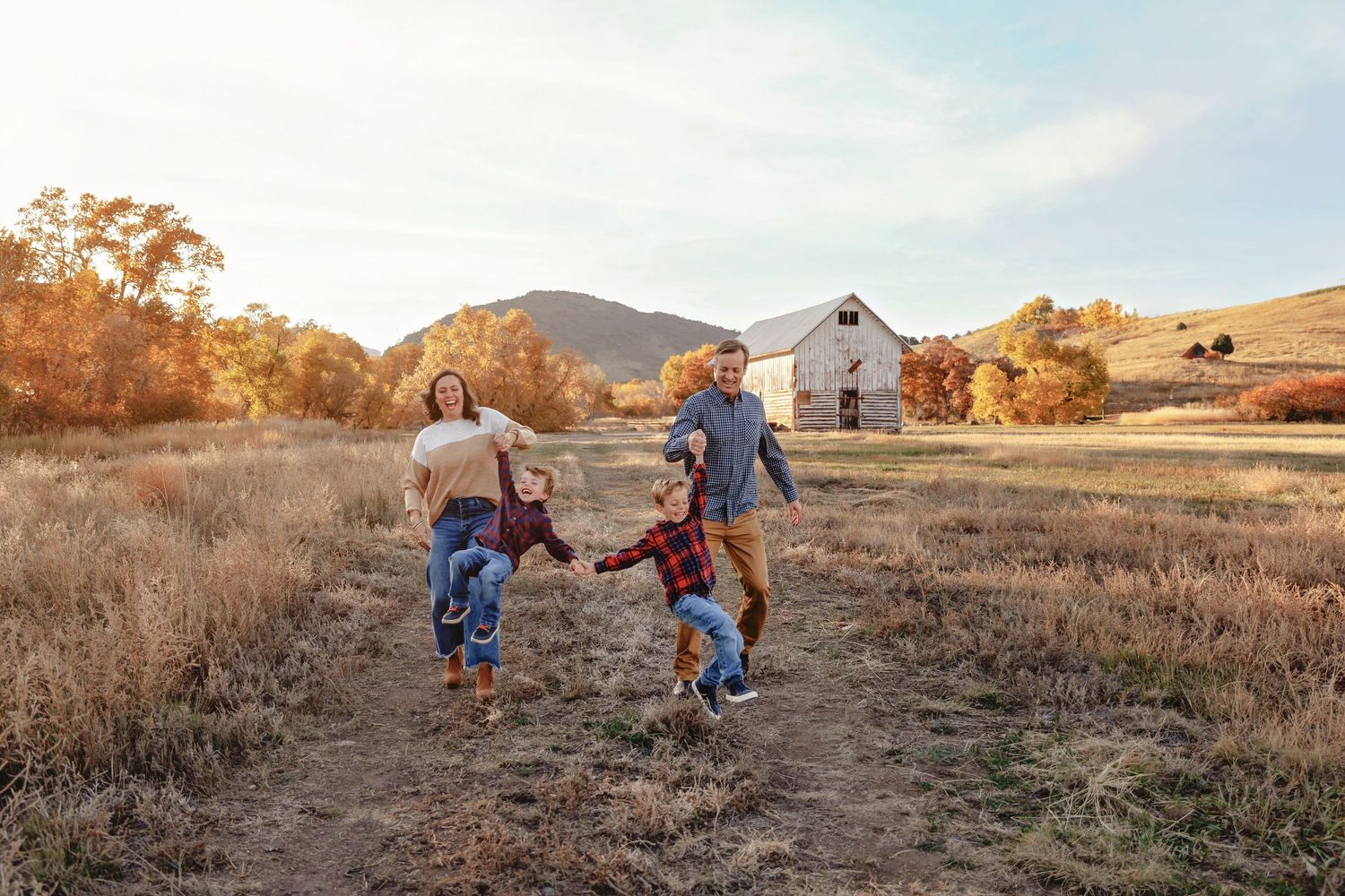 Family of four holding hands and walking through an autumn field with a white barn and mountains in background.