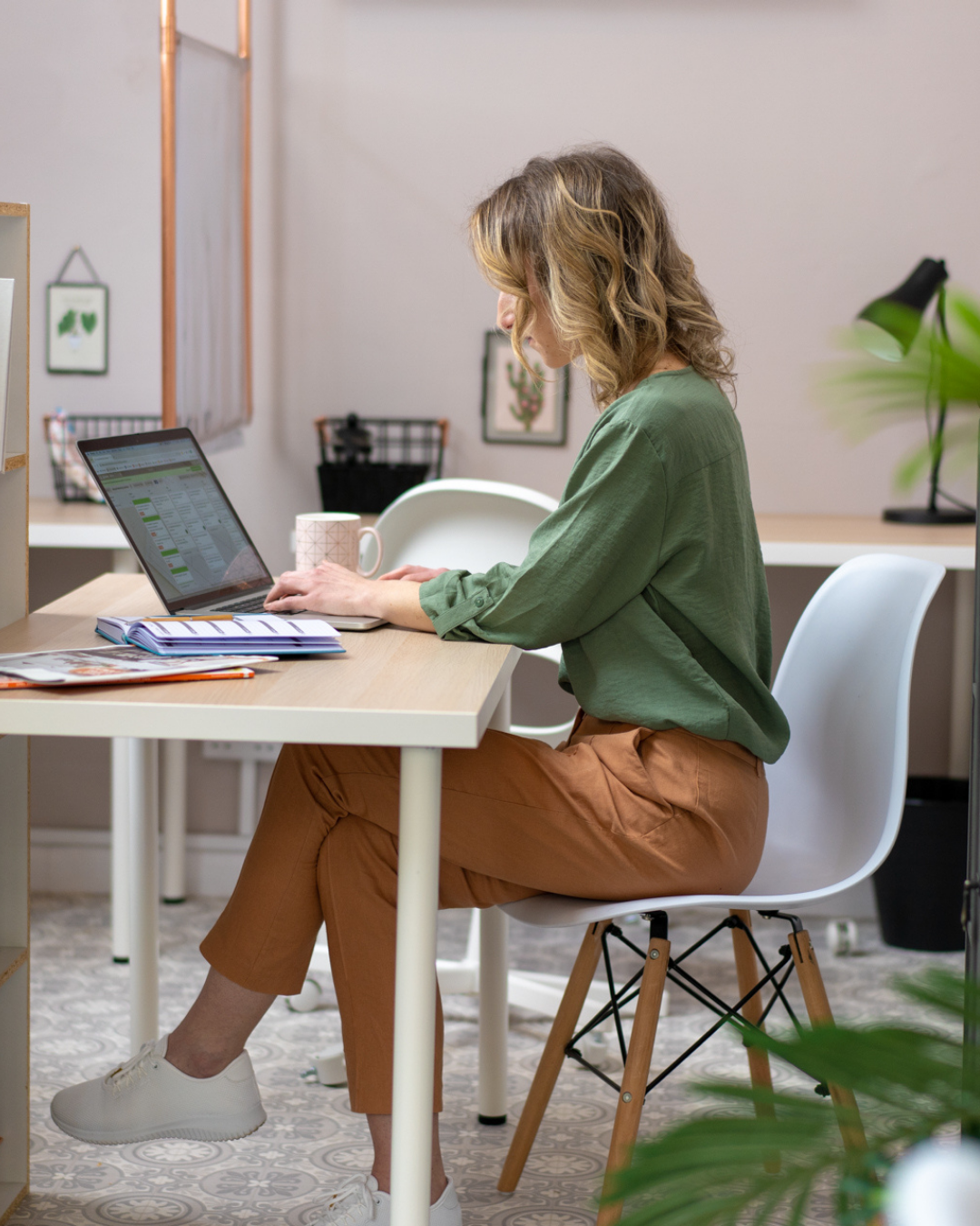 Woman in green shirt working on laptop at modern desk with plant decor.