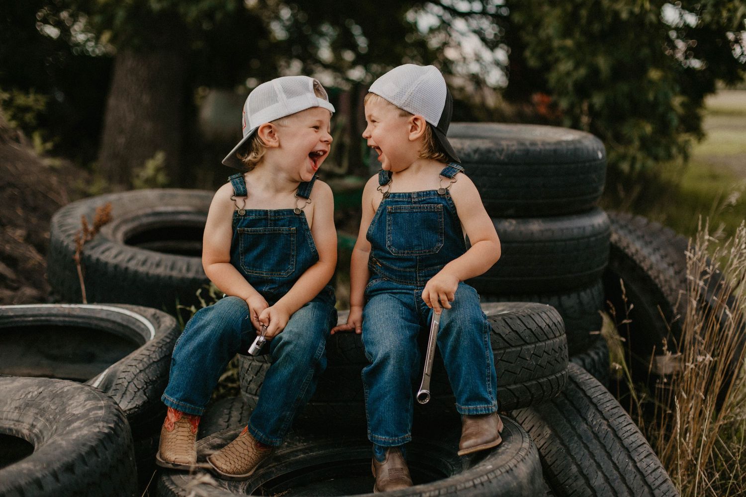 Two children in matching overalls laugh together while sitting on tractor tires.