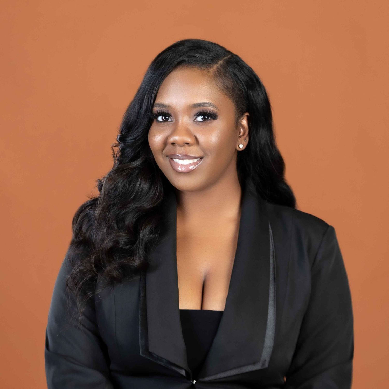 Professional portrait of person in black blazer with long dark wavy hair against orange background.