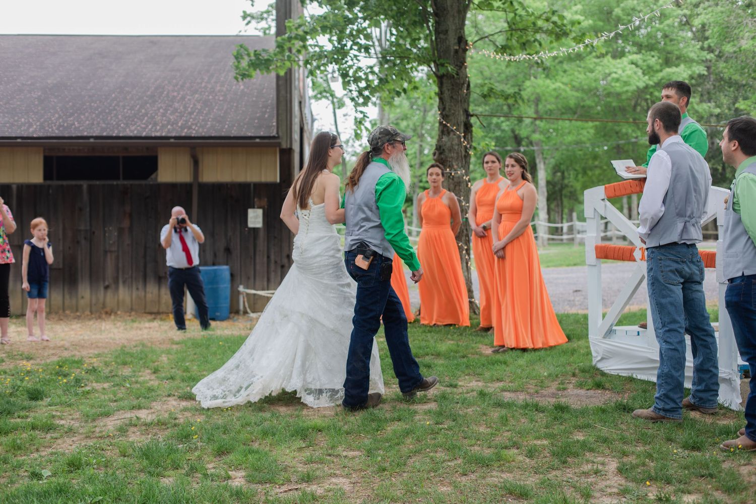 A wedding ceremony takes place outdoors with bridesmaids in bright orange dresses standing near a rustic building.