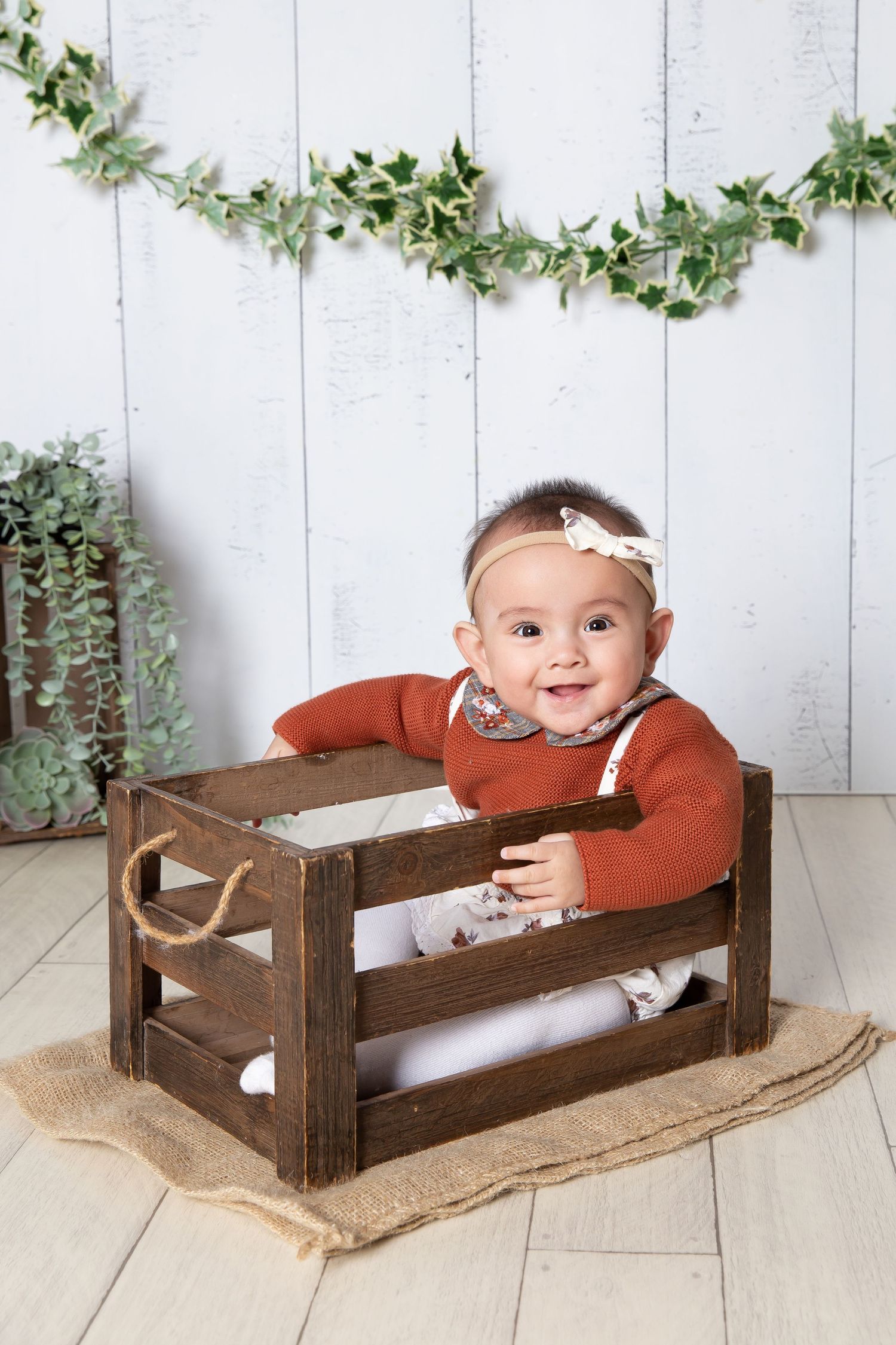baby sat in a crate on a white wooden background, wearing a brown top and headband