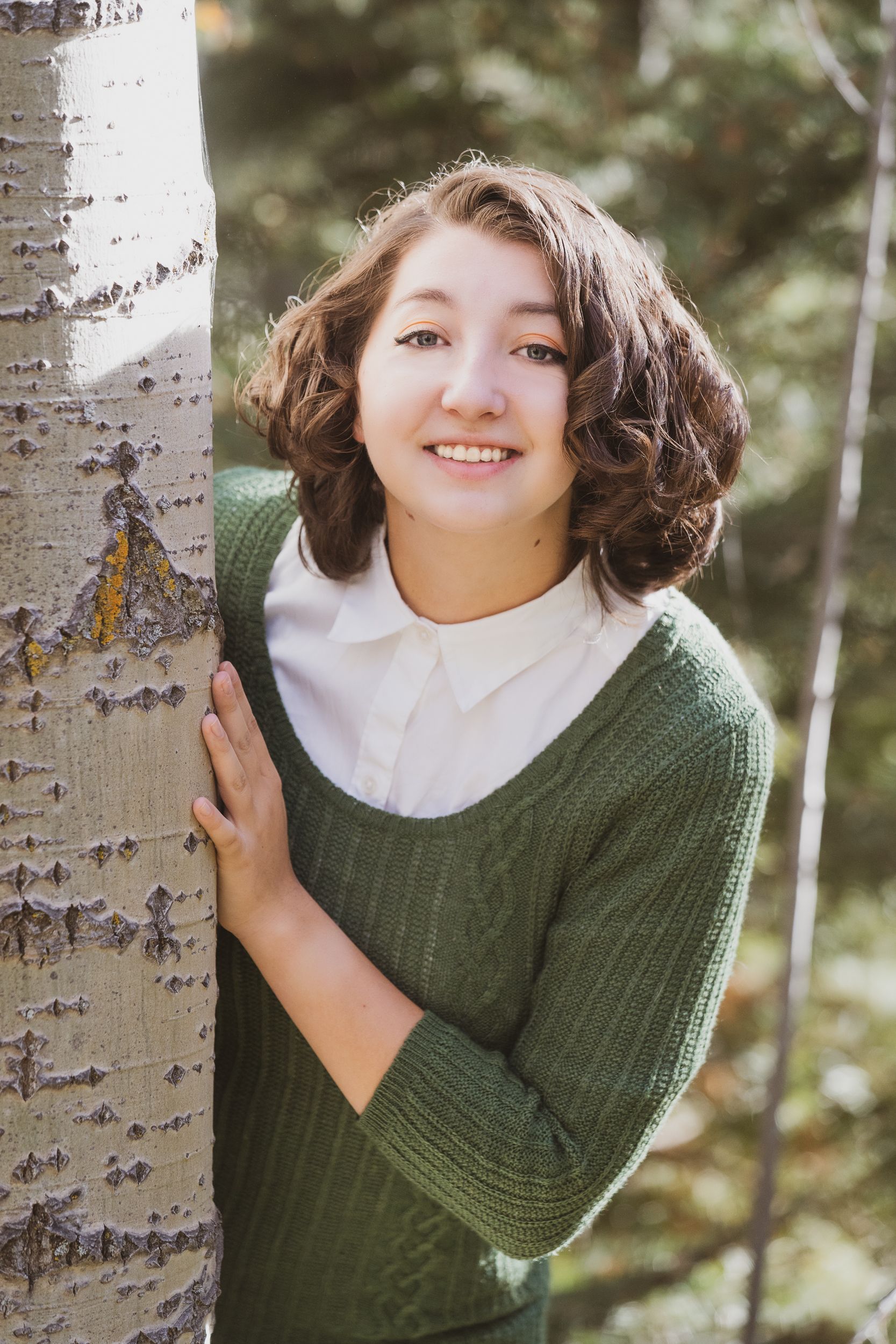 Portrait in nature showing someone in a green sweater leaning against a birch tree trunk.