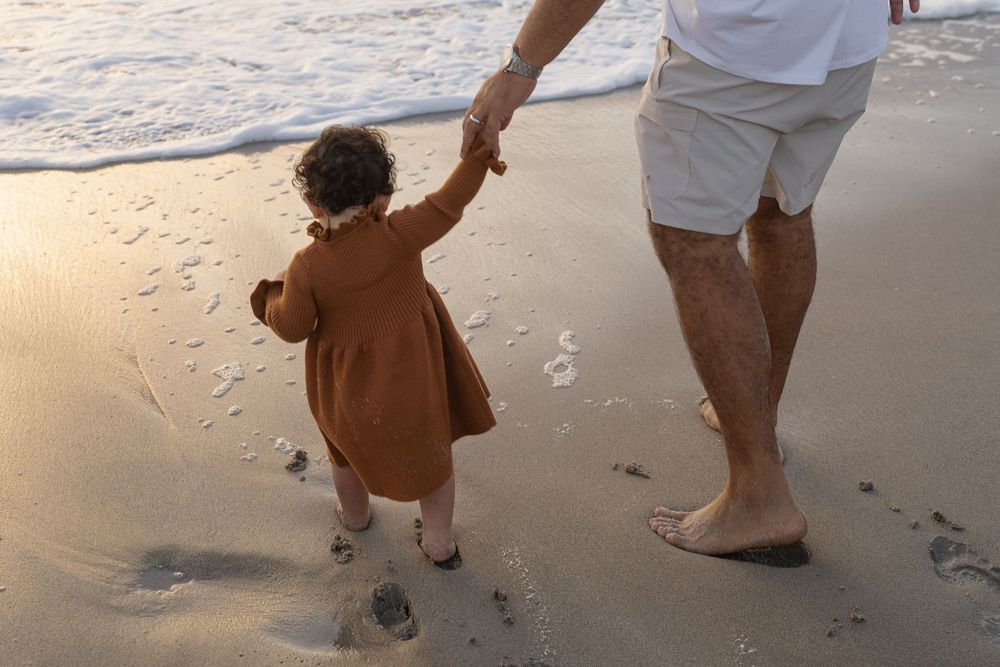 father holding his one year old daughters hand at the beach