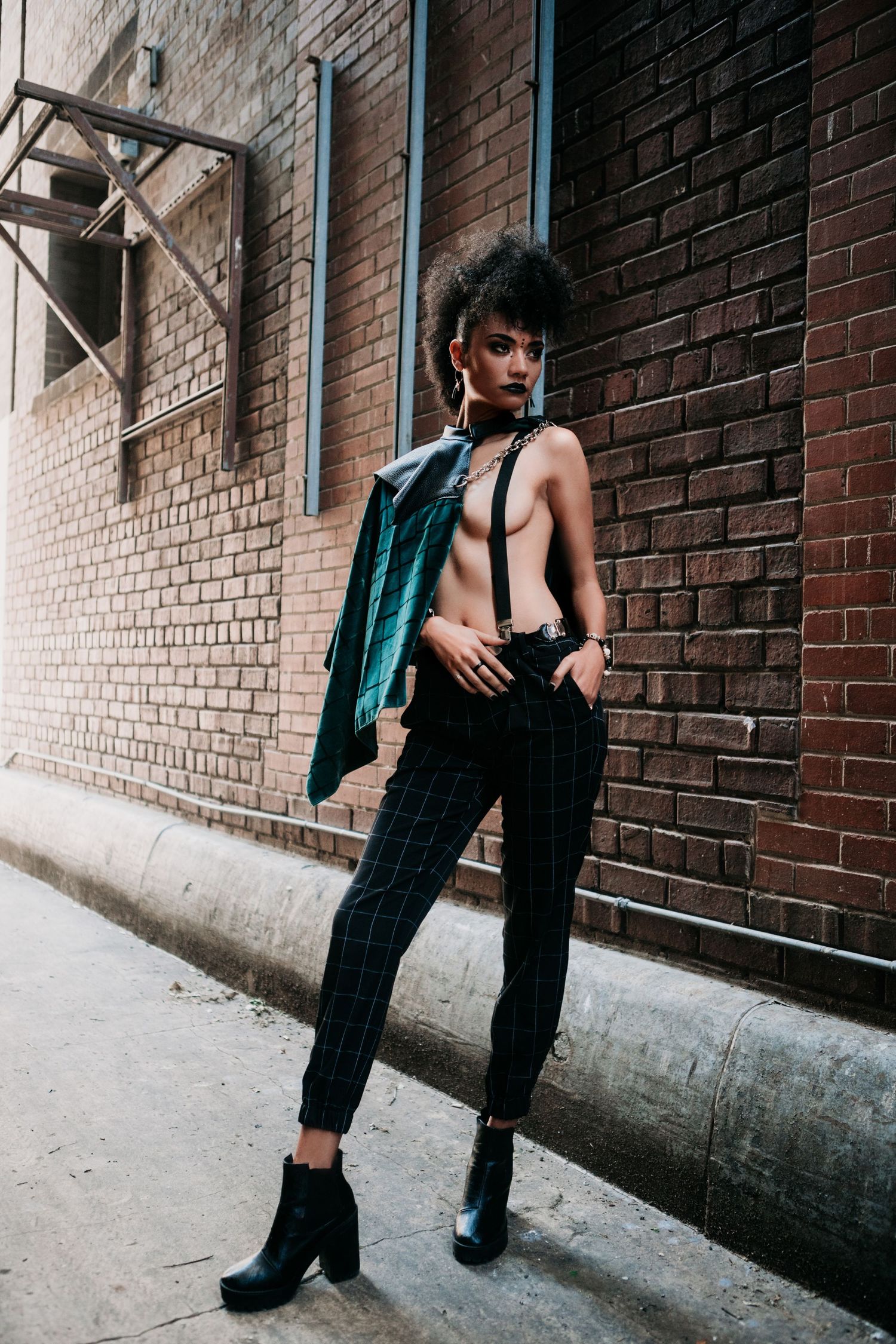 Fashion model posing against brick wall wearing teal vest, cropped top and plaid trousers with ankle boots.