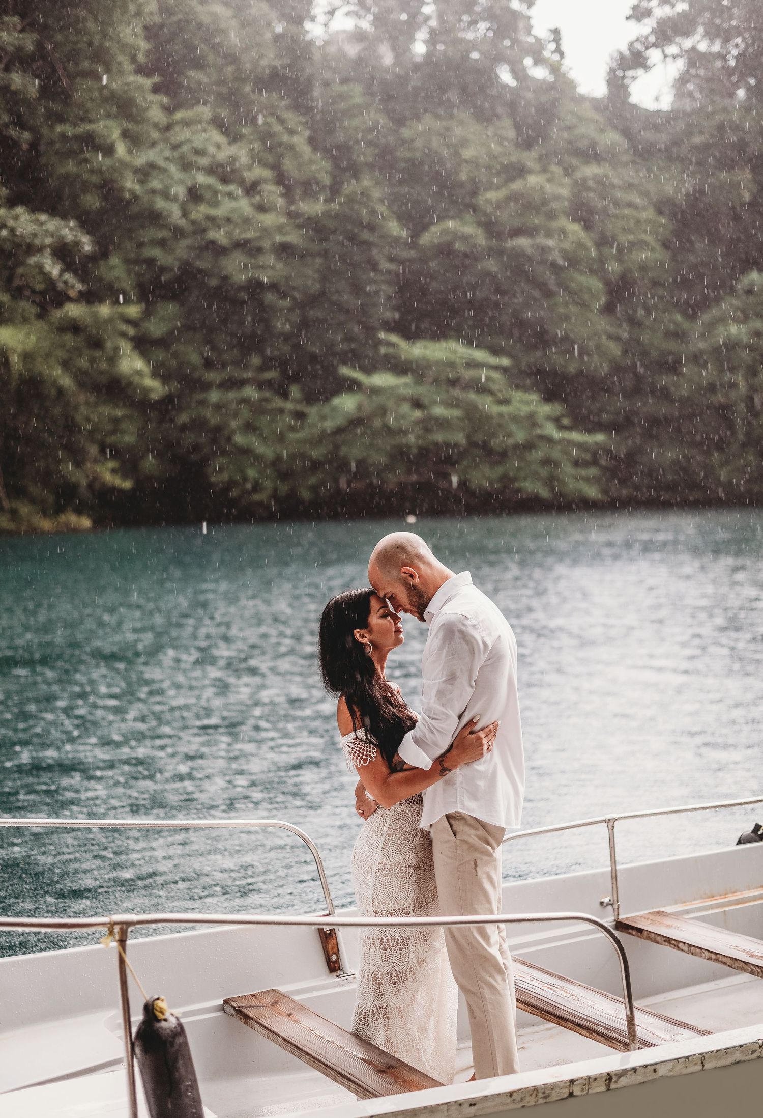 Romantic couple embracing on boat deck with turquoise lake and forest backdrop.