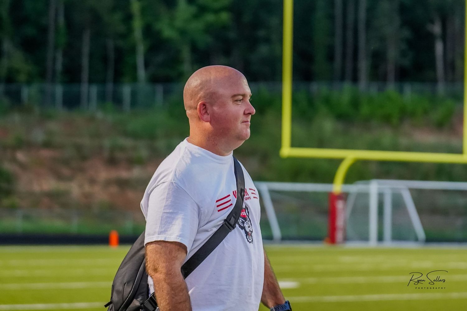 Person in white t-shirt with shoulder bag standing on athletic field with football goal post visible.