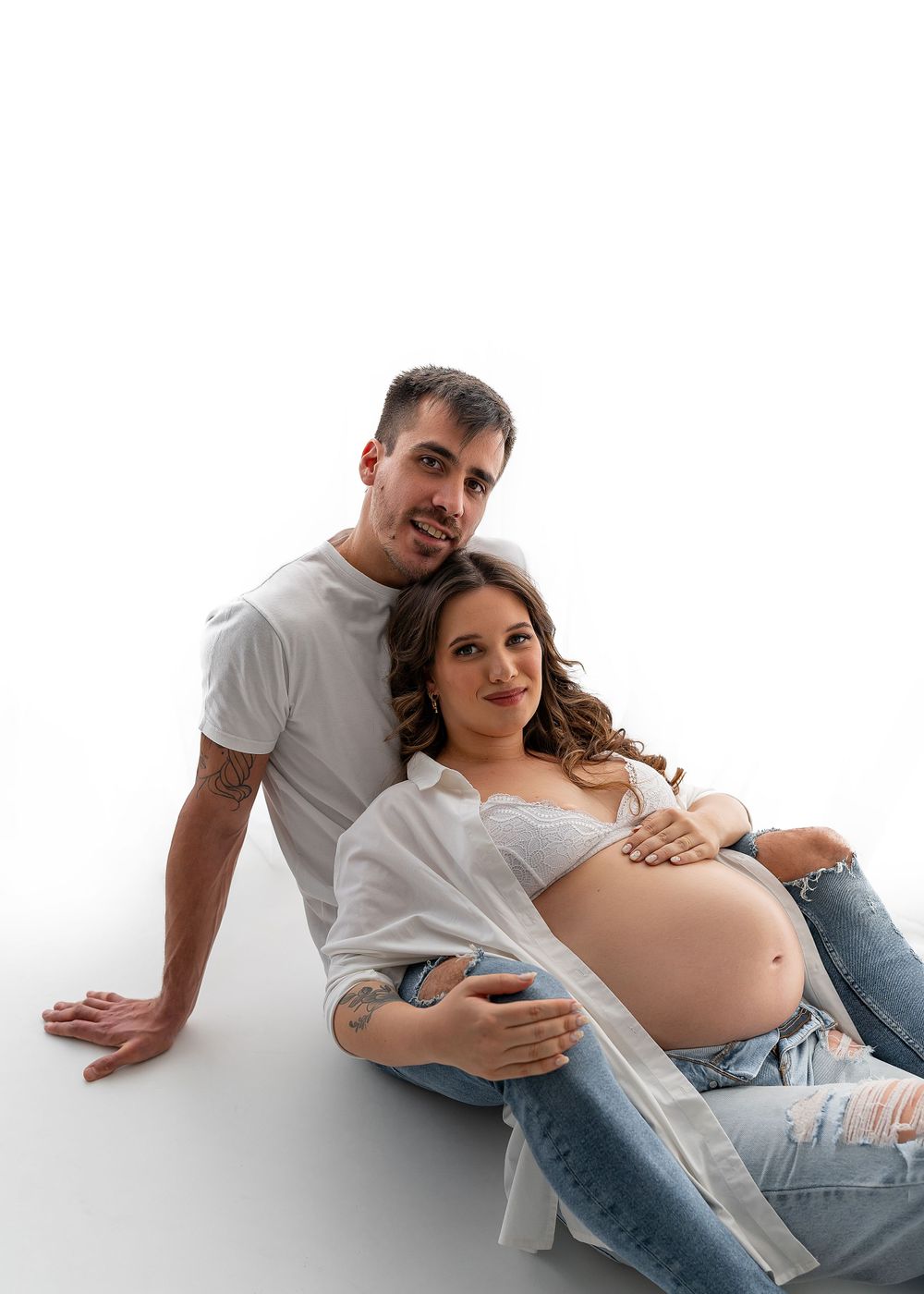 Couple's maternity photo with mother in white top and jeans resting on white floor.