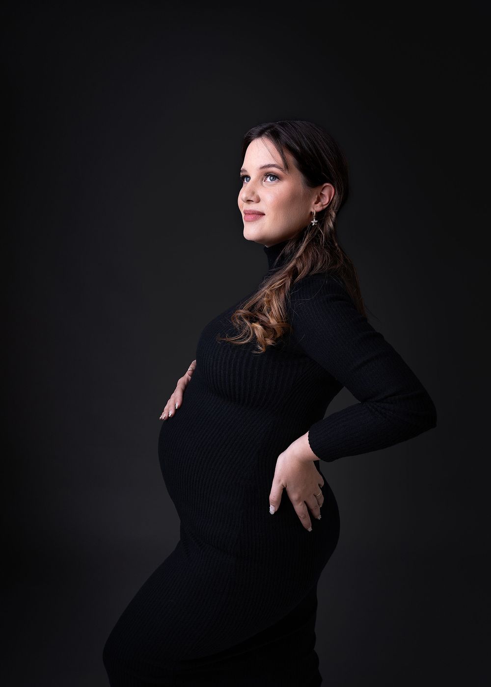 Maternity portrait of expectant mother in black turtleneck dress against dark background.