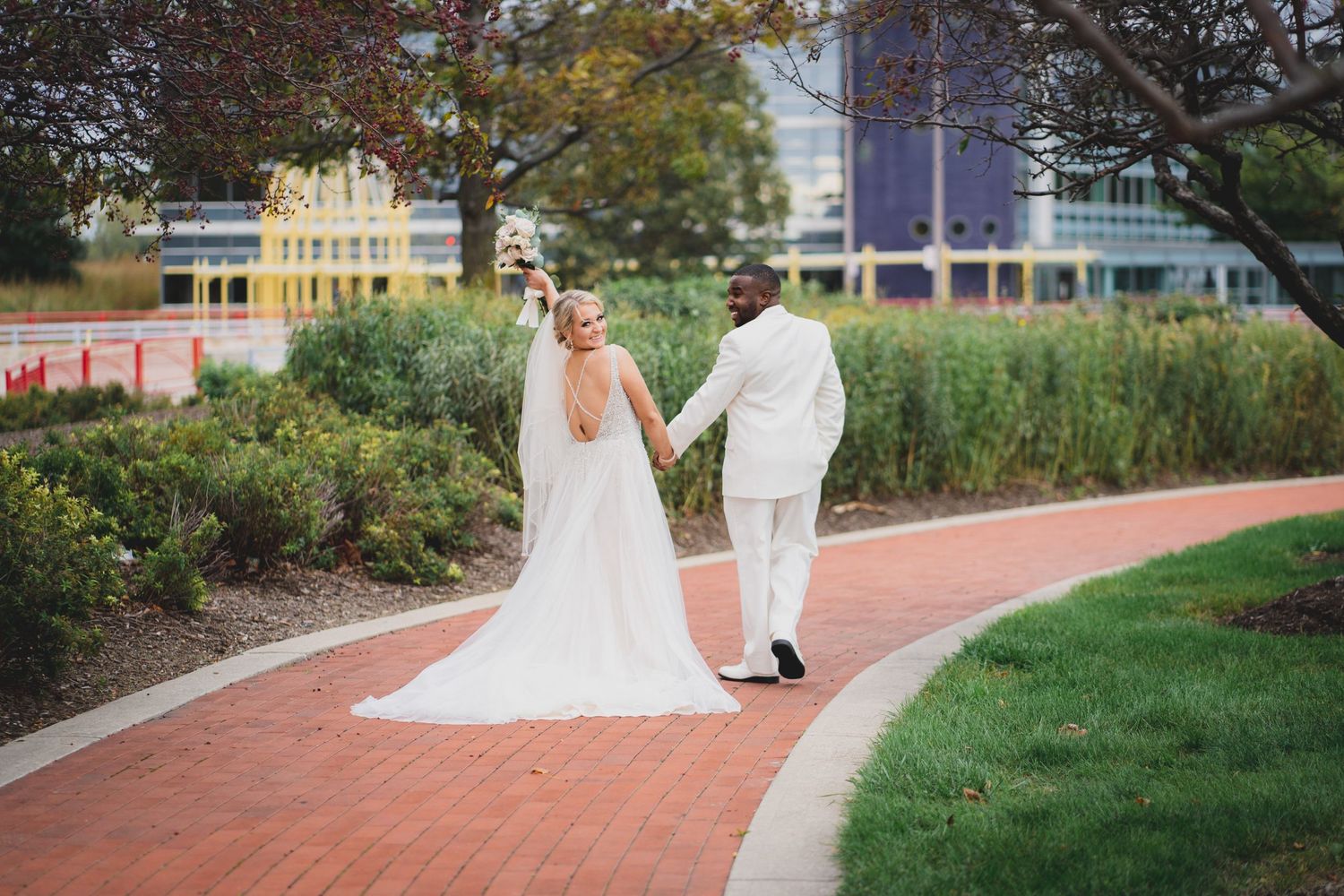 Couple in white attire walking along a curved brick pathway surrounded by landscaped gardens.