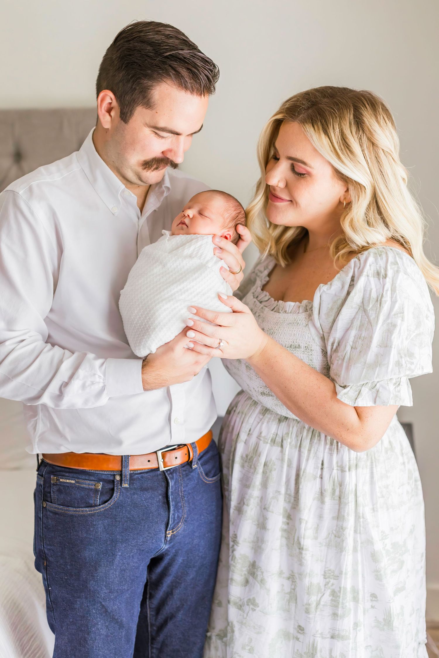 Parents in casual attire holding their newborn baby wrapped in white.