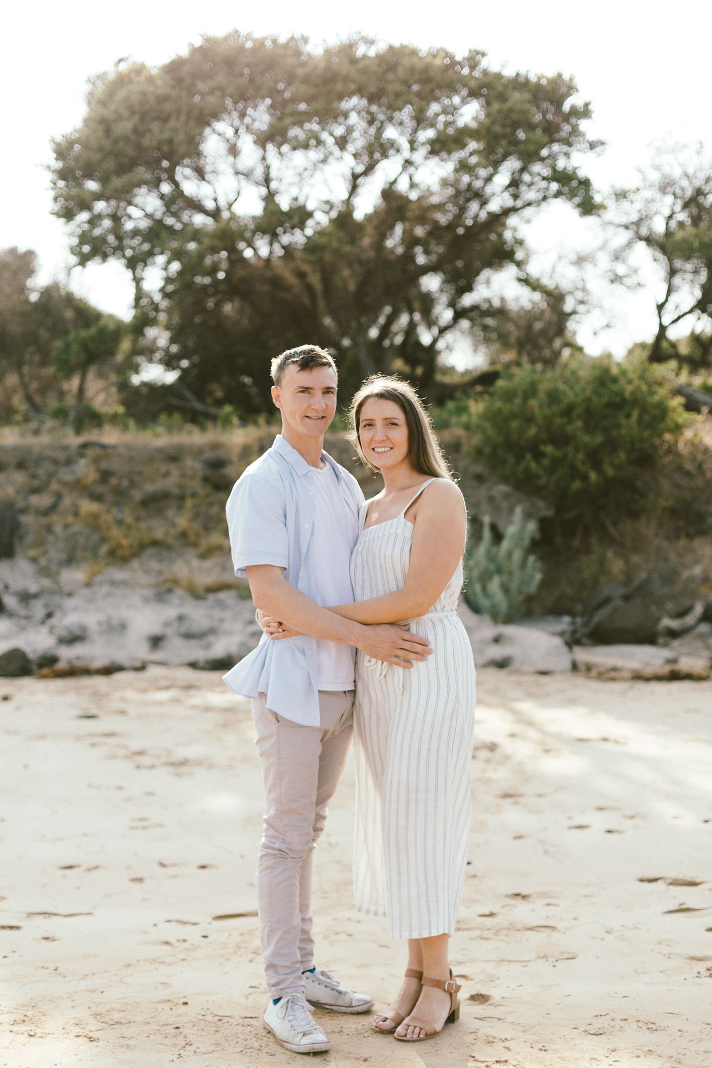 A couple in white and light blue outfits embraces on a sandy beach.