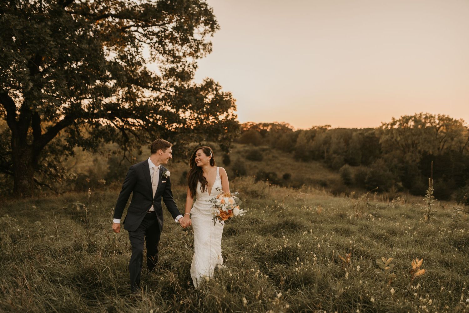 A newlywed couple walks through a golden field during sunset in their wedding attire.