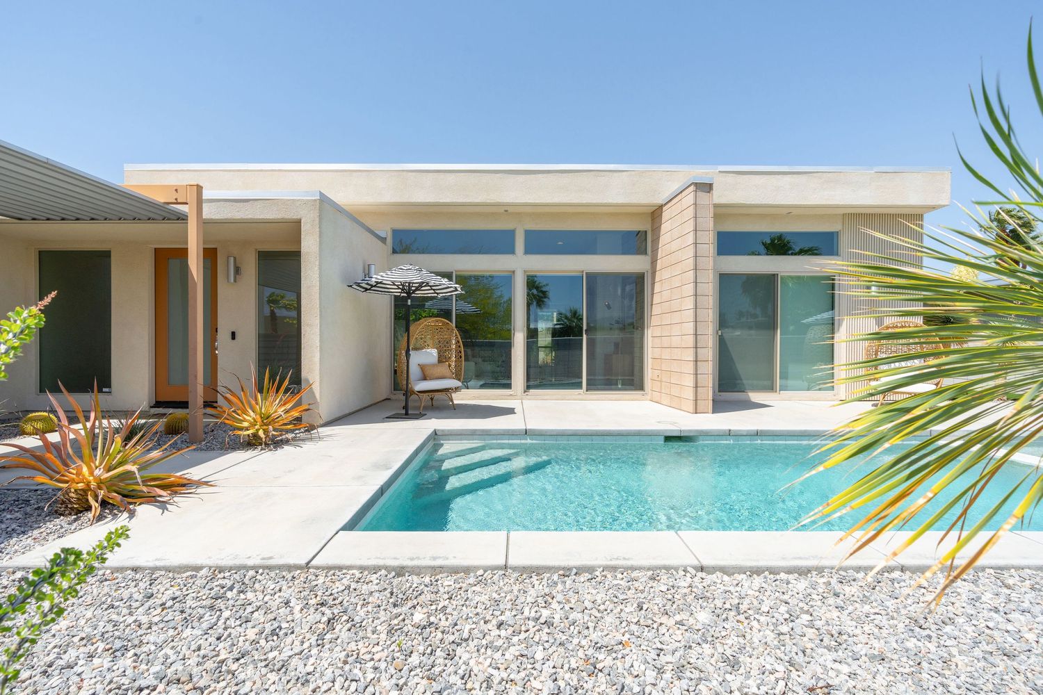 Modern desert house exterior with sliding glass doors, swimming pool, and minimalist landscaping in sunlit setting.