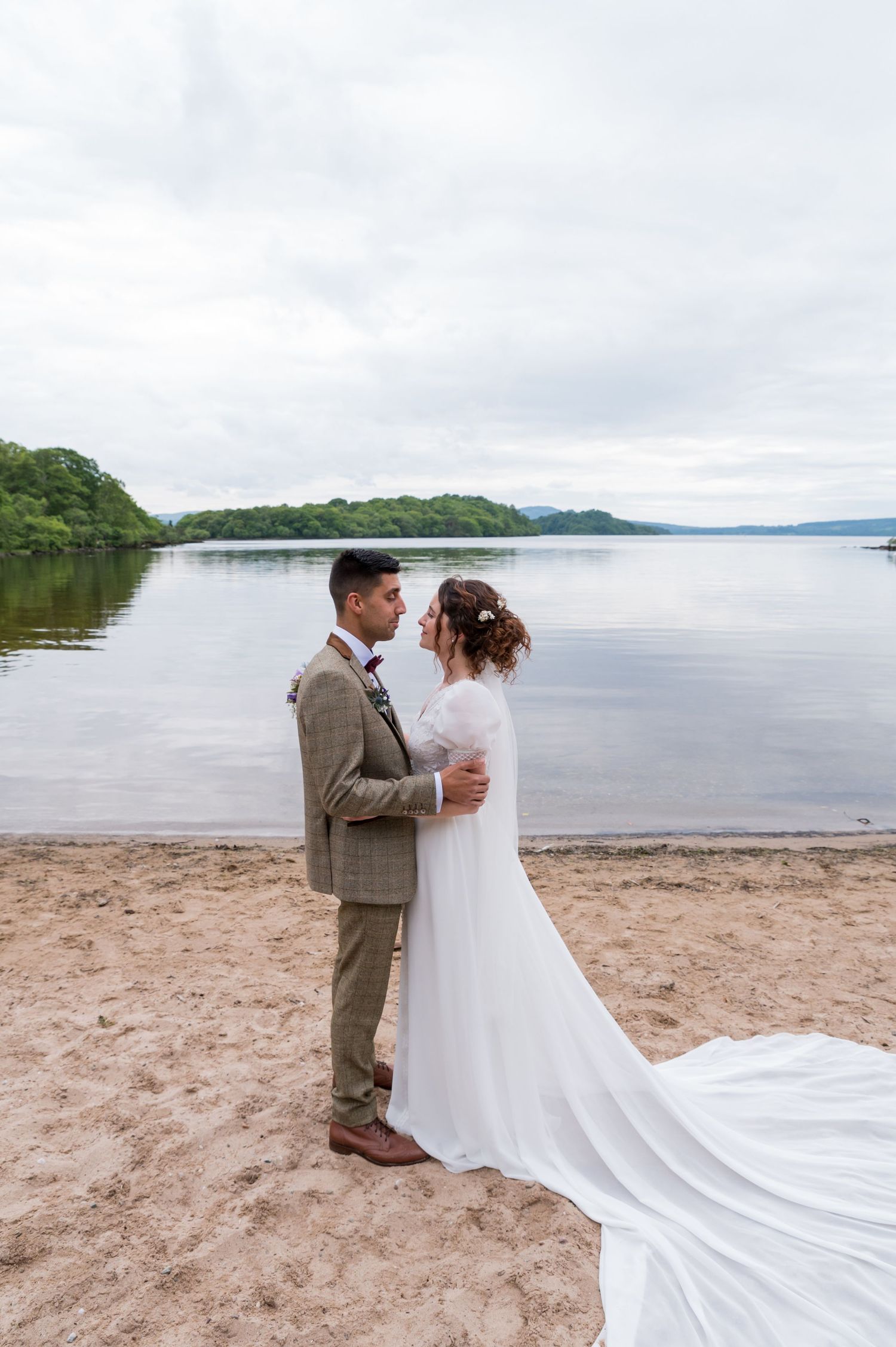A wedding couple embraces on a beach shore with a serene lake and hills in the background.