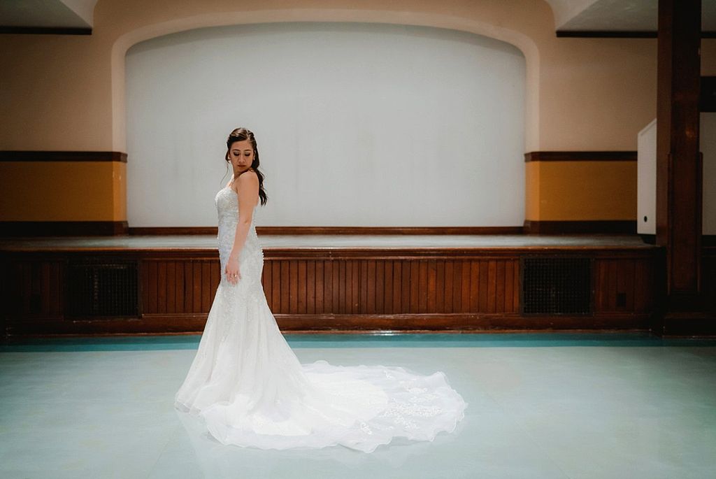 A bride in a flowing white mermaid wedding gown stands elegantly in a room with wooden paneling and mint green floors.