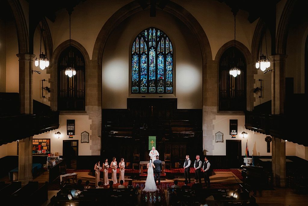 Church wedding ceremony under gothic arches with stained glass windows and pendant lighting.