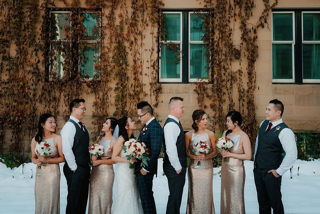 Bridal party in formal attire poses against an ivy-covered building in winter.