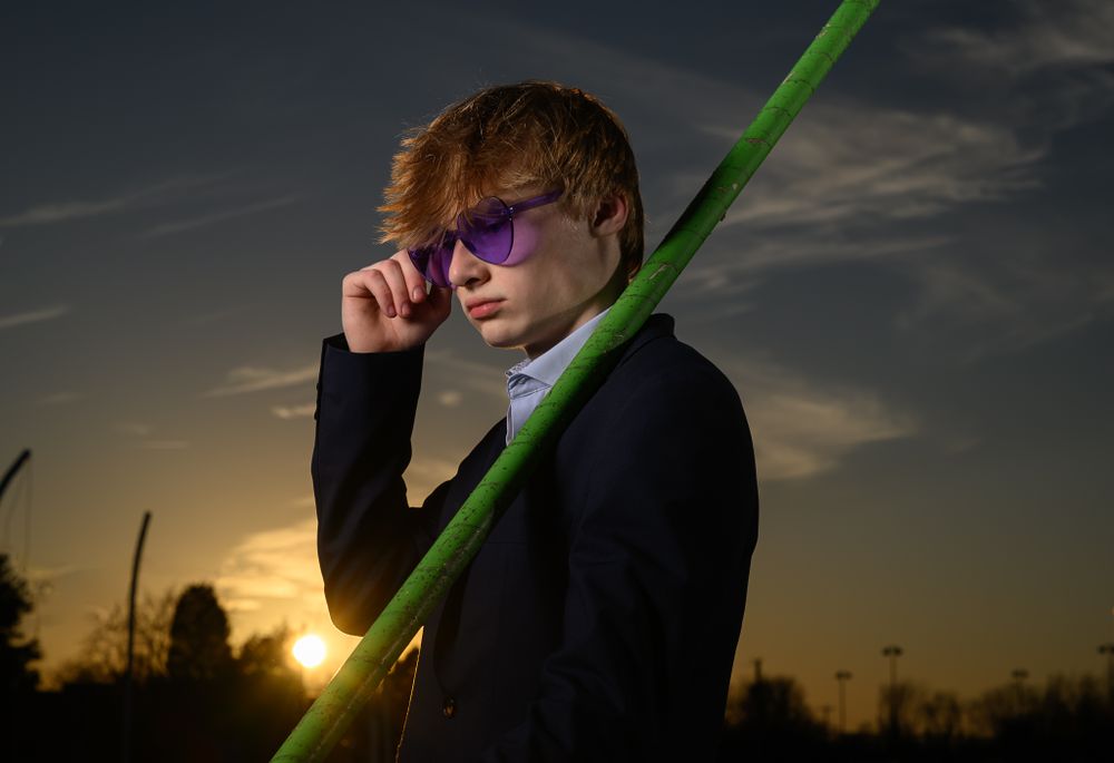 Figure in dark clothing holds green pole against sunset sky with stadium lights visible.