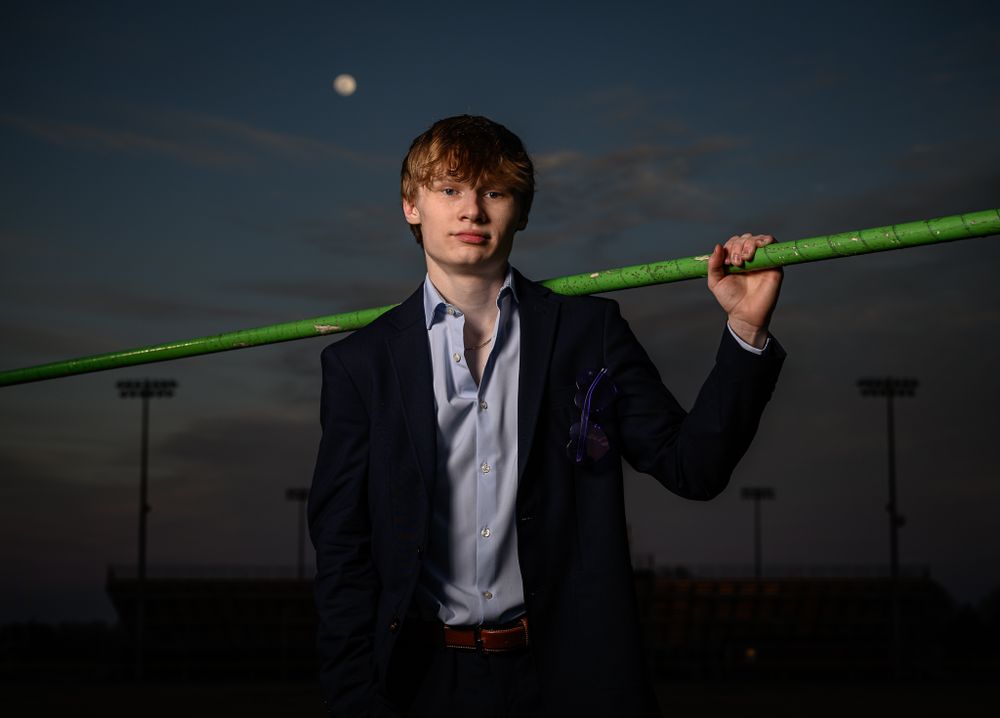 Person in formal suit poses with green pole at dusk with moon and stadium lights in background.