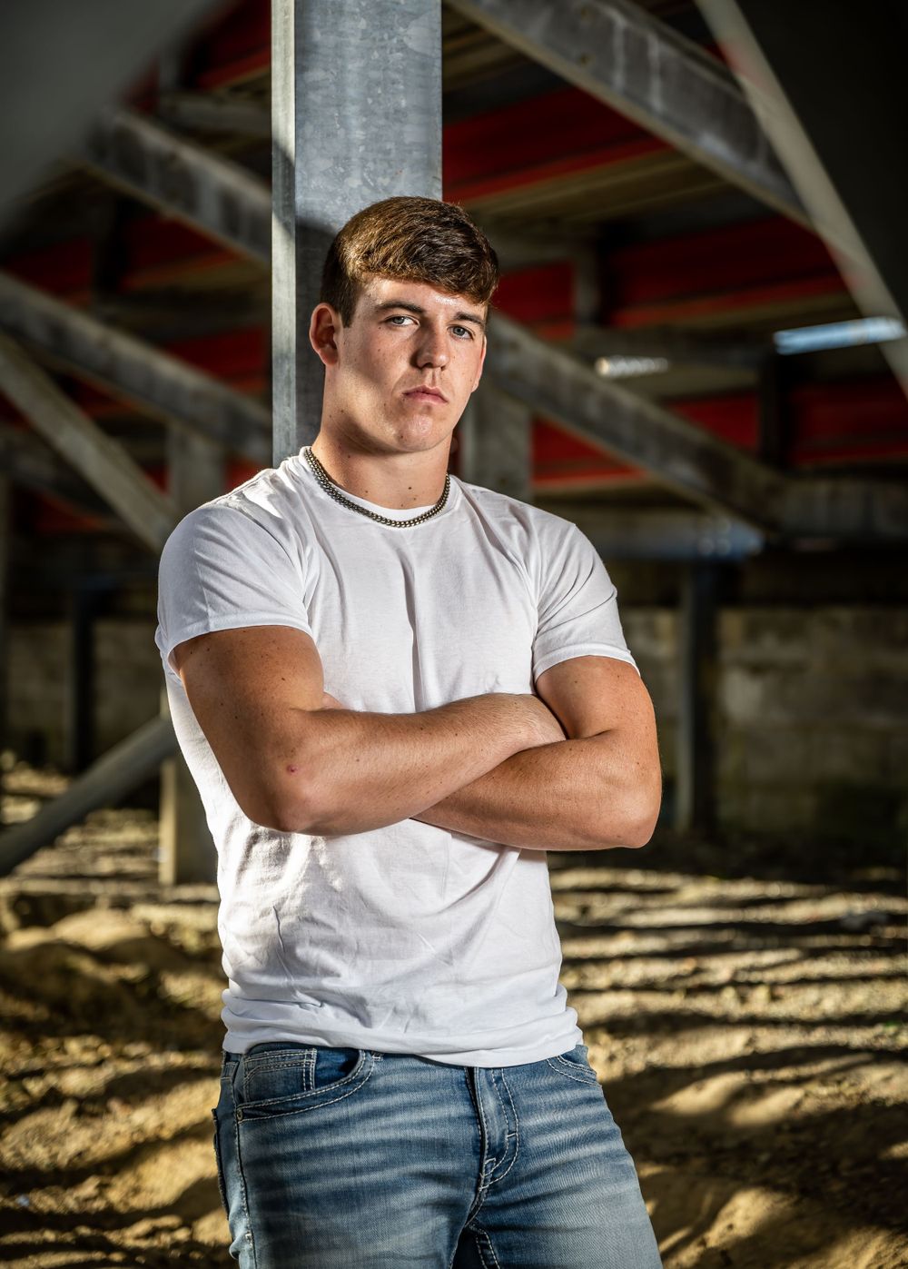 Athletic figure in white t-shirt poses with crossed arms under industrial architecture.