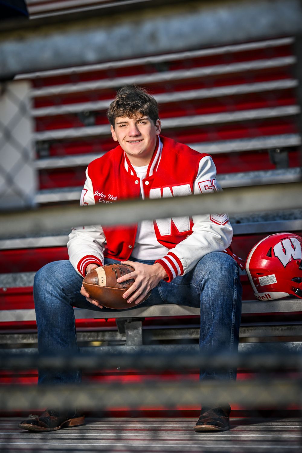 Athletic figure in varsity jacket poses with football on bleachers with American flag backdrop.