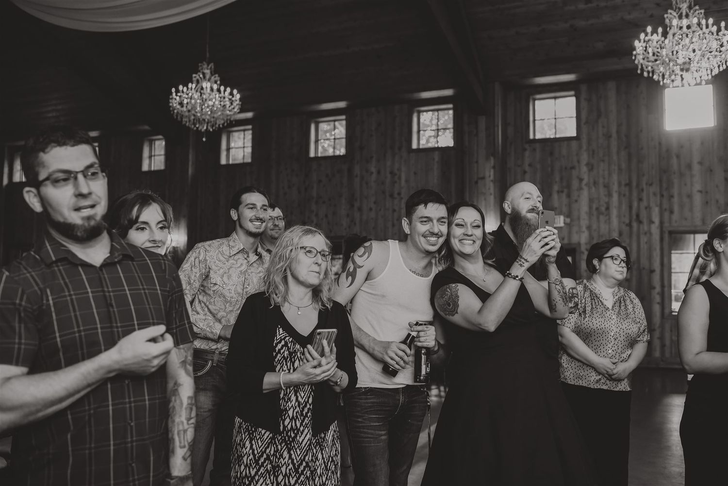 Wedding guests watch and smile during a reception in an elegant wood-paneled room with chandeliers.