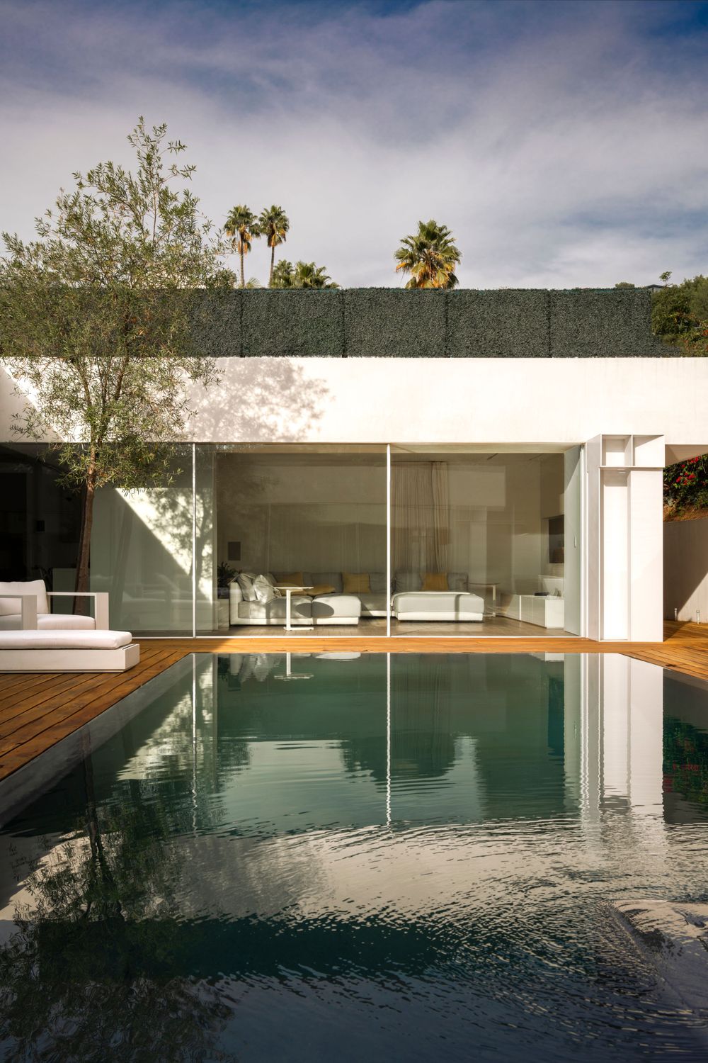 Modern poolside patio with wooden deck and glass walls reflecting the afternoon sun.