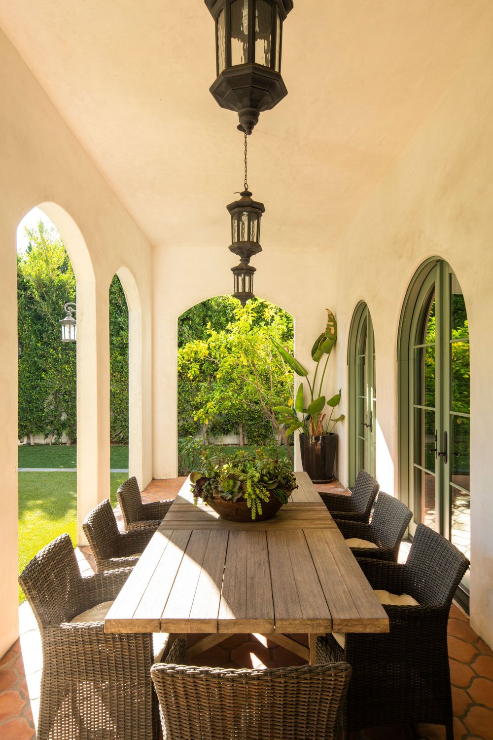 Mediterranean-style covered porch with wicker dining chairs and wooden table under lantern lights.